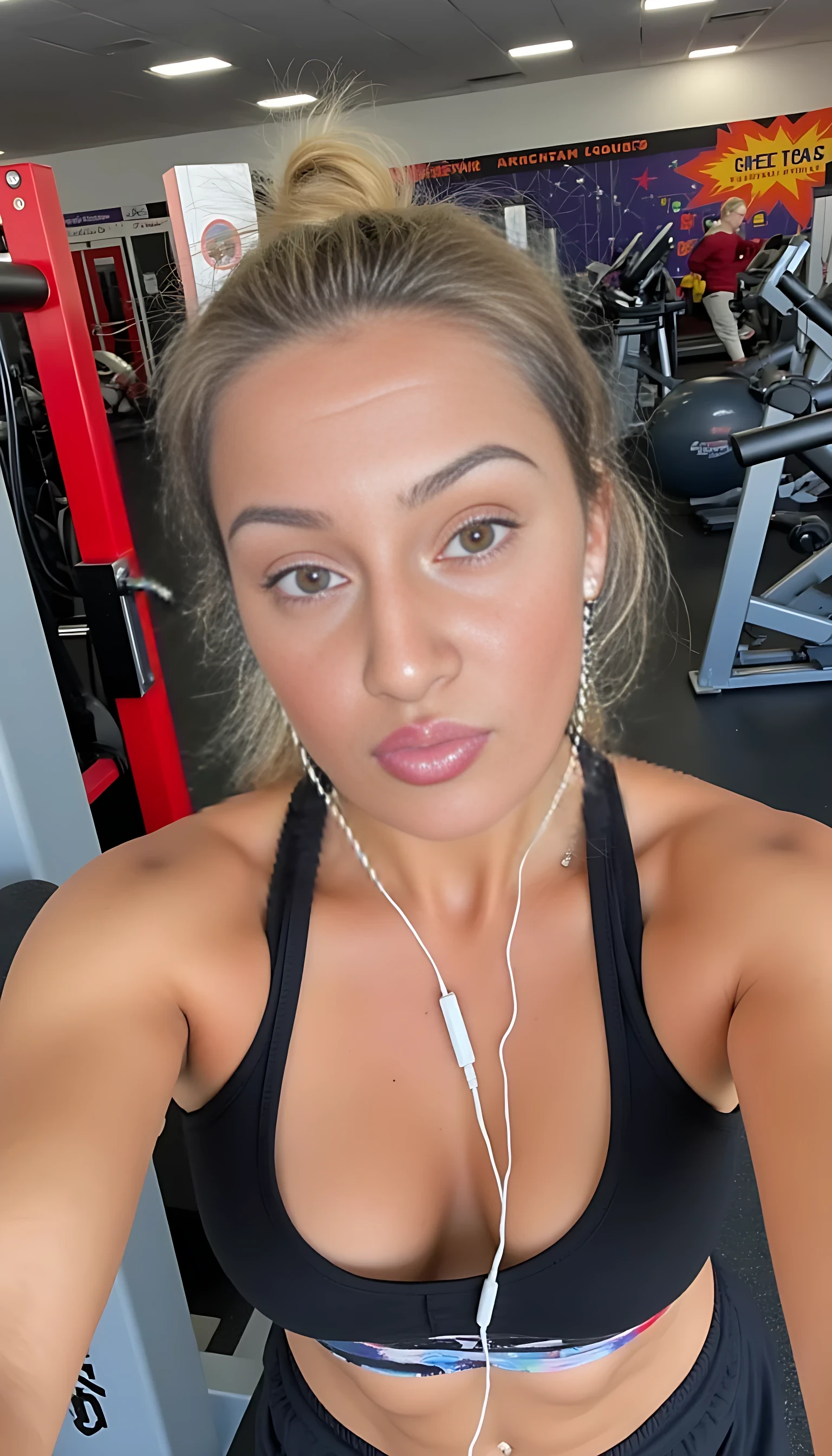 A woman takes a selfie in a gym environment. She is wearing workout attire, including a sports bra, and has earphones in. Her expression is playful, with raised eyebrows and puckered lips. The gym setting includes exercise equipment like treadmills and stationary machines in the background, with a vibrant and lively atmosphere.