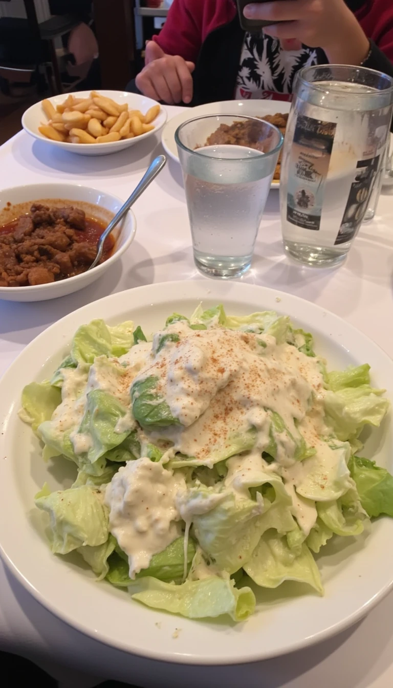 boring low quality snapchat photo circa 2015, this picture is being taken in a restaurant. The picture is of large caesar salad with a glass of water next to it. There are other food dishes and drink glasses on the table as well.  All the food dishes are white.