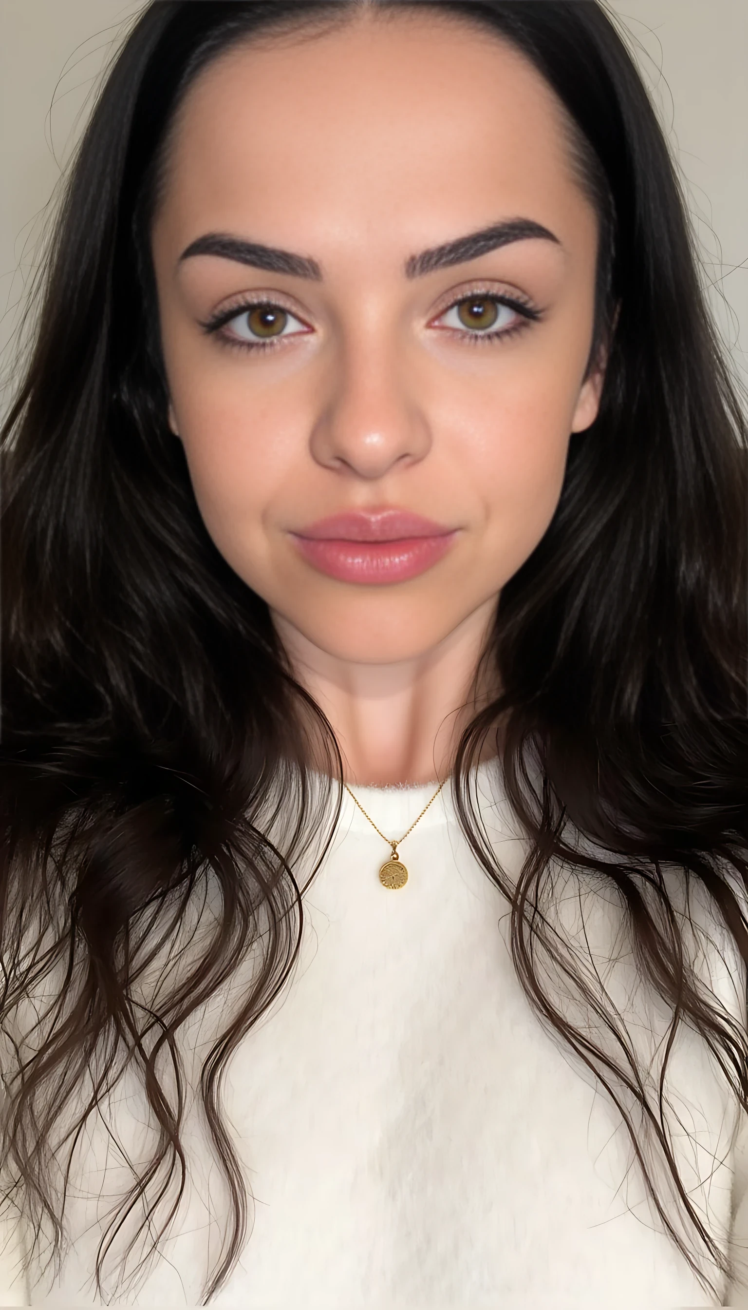 photo of Lena medium Close-up portrait of a beautiful young woman with fair skin and smooth complexion, soft natural lighting. She has long, dark brown wavy hair, well-groomed thick eyebrows, almond-shaped hazel-green eyes with defined eyelashes, and subtle winged eyeliner. Her lips are full with a natural rosy tone, and she wears a soft matte nude lipstick. Minimal makeup look with a focus on natural beauty. She has a symmetrical face, slightly angled jawline, and a calm, confident expression. She is wearing a white fluffy sweater and a small golden pendant necklace. Neutral background, studio-quality photograph, photorealistic detail, 85mm lens, shallow depth of field.
