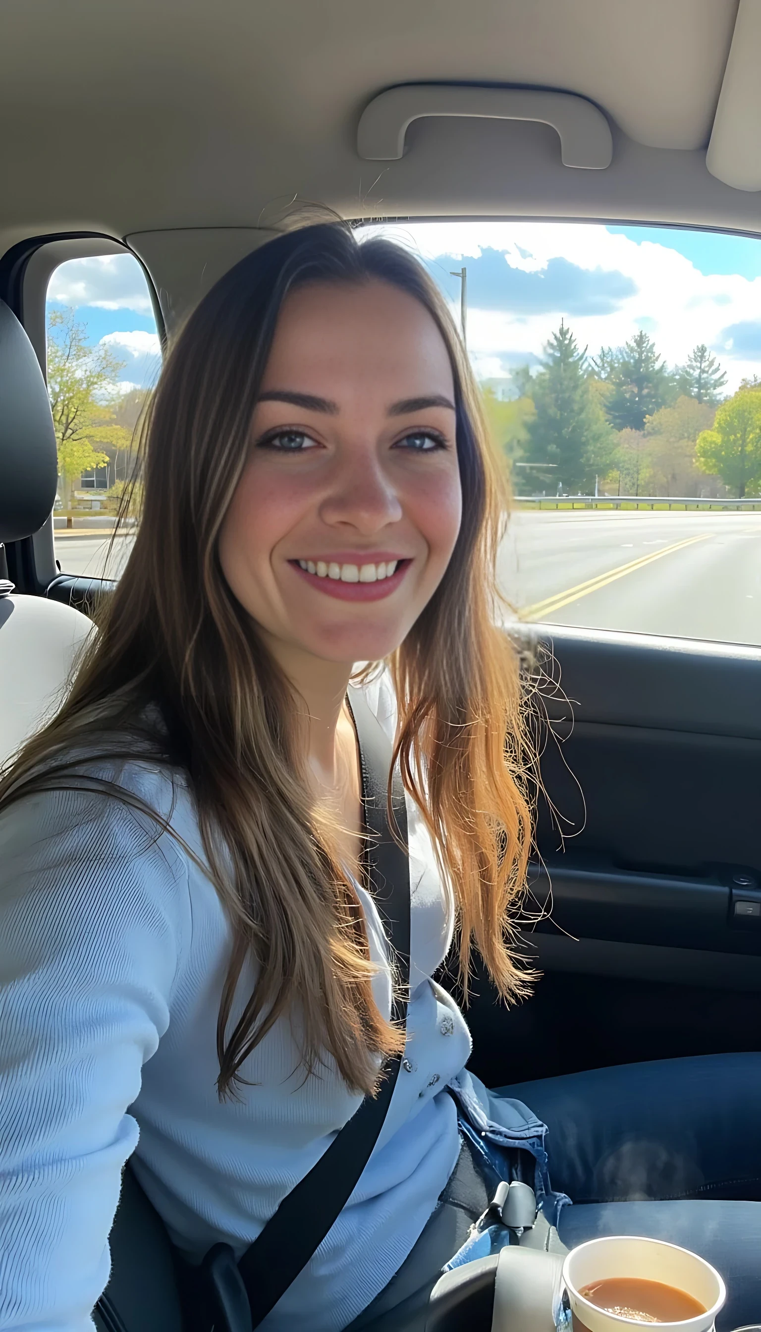 boring low quality snapchat photo circa 2015, A young woman with a warm smile, sitting in the driver's seat of a cozy car. She has soft, wavy brown hair cascading past her shoulders. The sunlight streams through the window, casting a gentle glow on her face. The interior of the car is neat and comfortable, with a coffee cup in the cup holder and a small plant on the dashboard. The background outside the window shows a scenic road lined with trees and blue skies