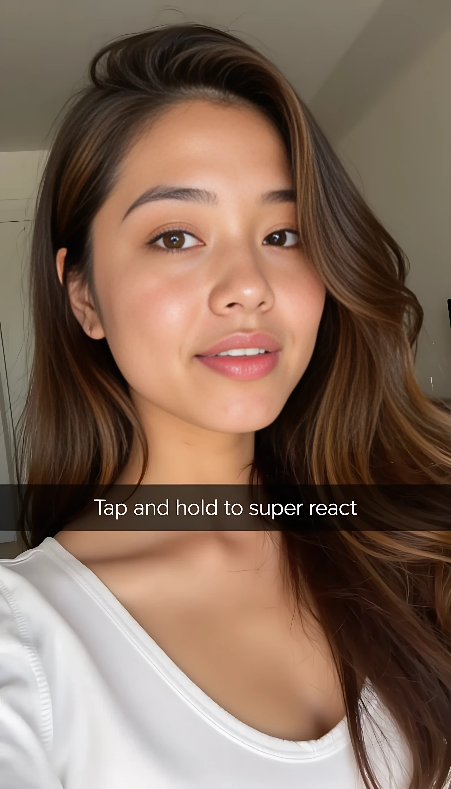 A photograph of a young woman with long, wavy brown hair, fair skin, and full lips. She's wearing a white top, with a blurred background. A text overlay reads "Tap and hold to super react."