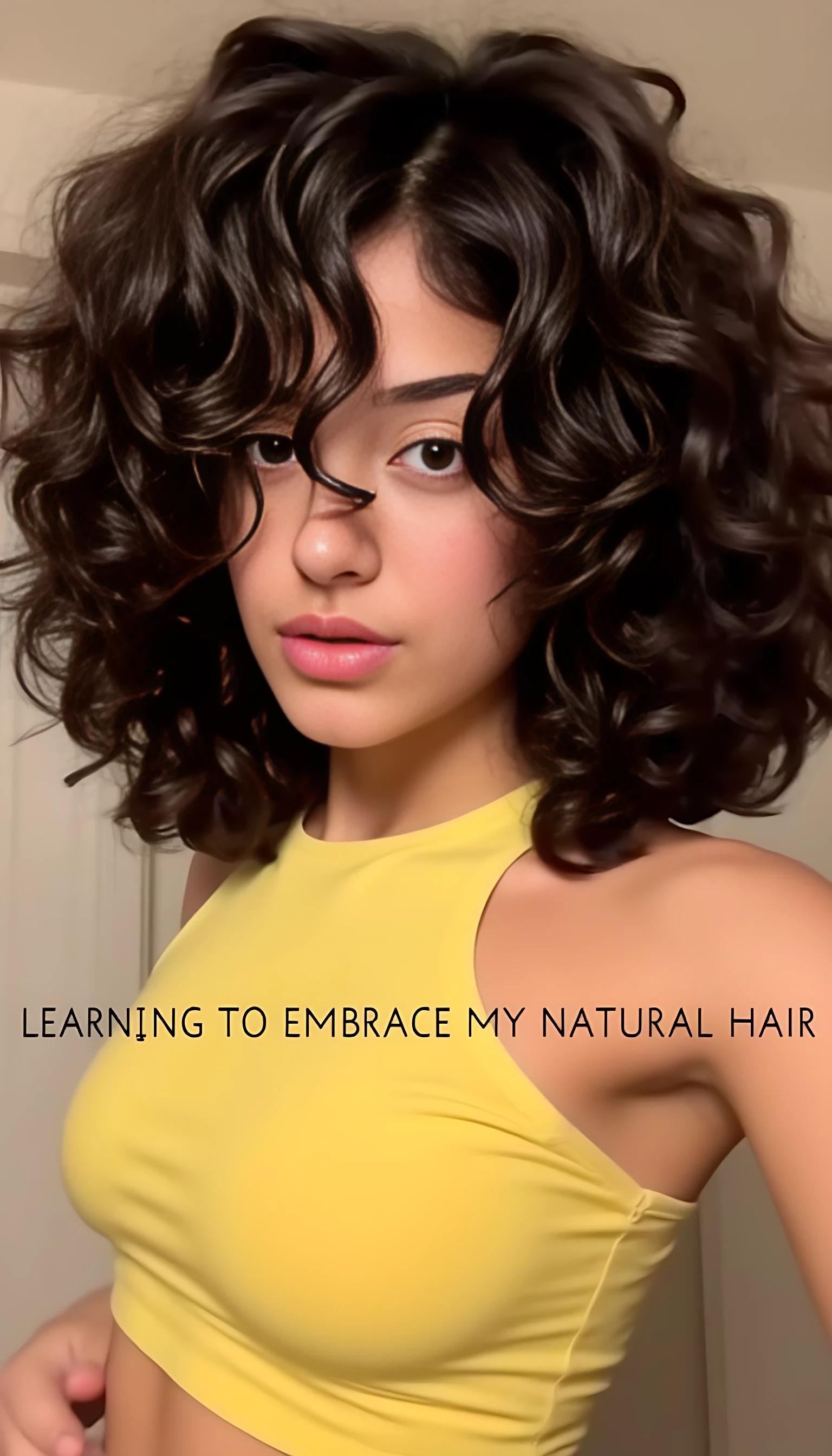 A confident person standing indoors with voluminous, curly dark hair, wearing a yellow crop top. The lighting creates a warm and inviting atmosphere, enhancing the person's natural features. The text "LEARNING TO EMBRACE MY NATURAL HAIR" is displayed across the image. The background shows a simple, modern room with soft shadows and neutral tones.