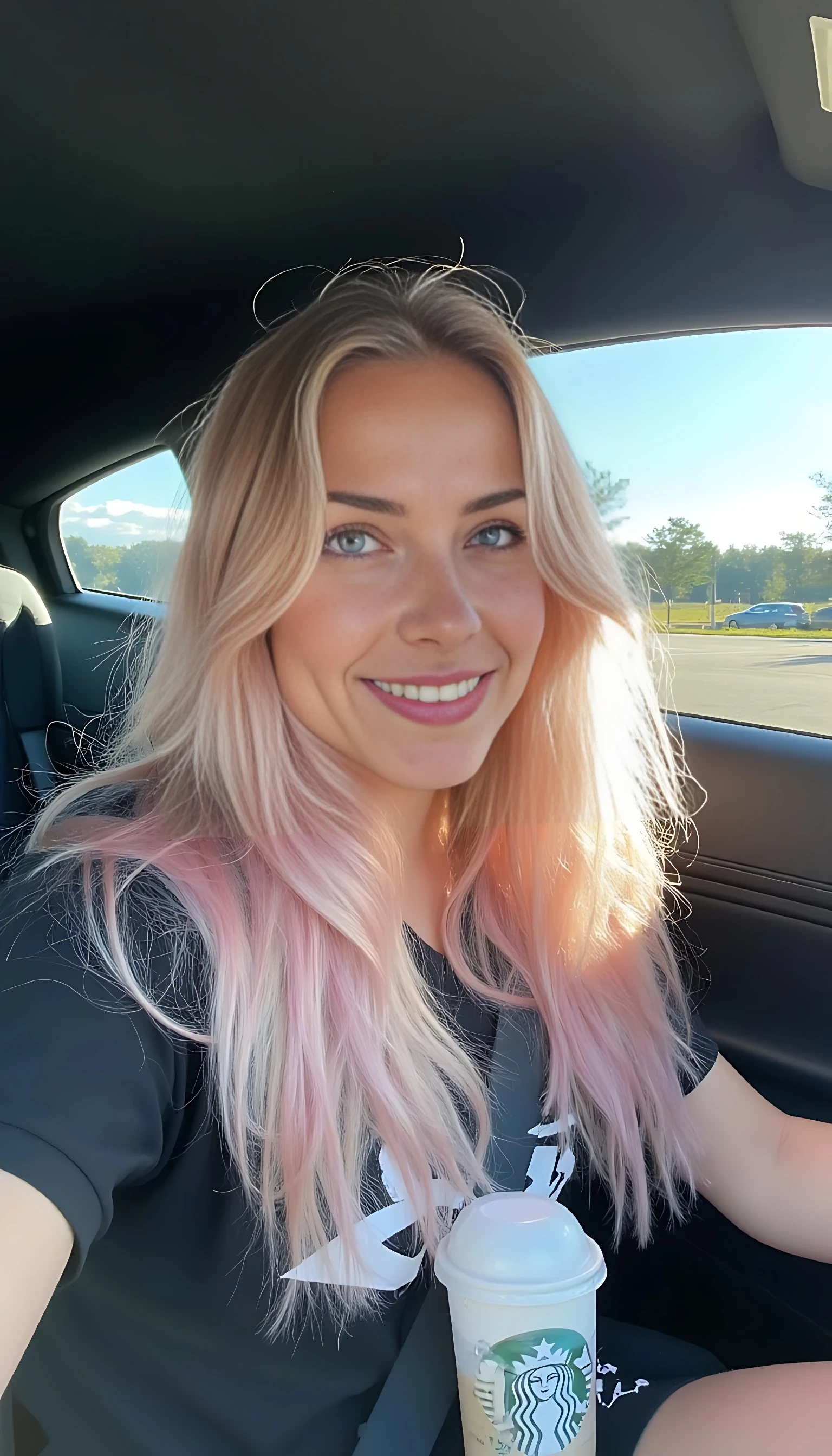 boring low quality snapchat photo circa 2015, A young woman, warm smile, sitting in the driver's seat of a Ferrari  car. She has soft, wavy light pink hair cascading past her shoulders. The sunlight streams through the window, casting a gentle glow on her face. The interior of the car is neat and comfortable, with a Starbucks refresher in the cup holder. The background outside the window shows a scenic road lined with trees and blue skies