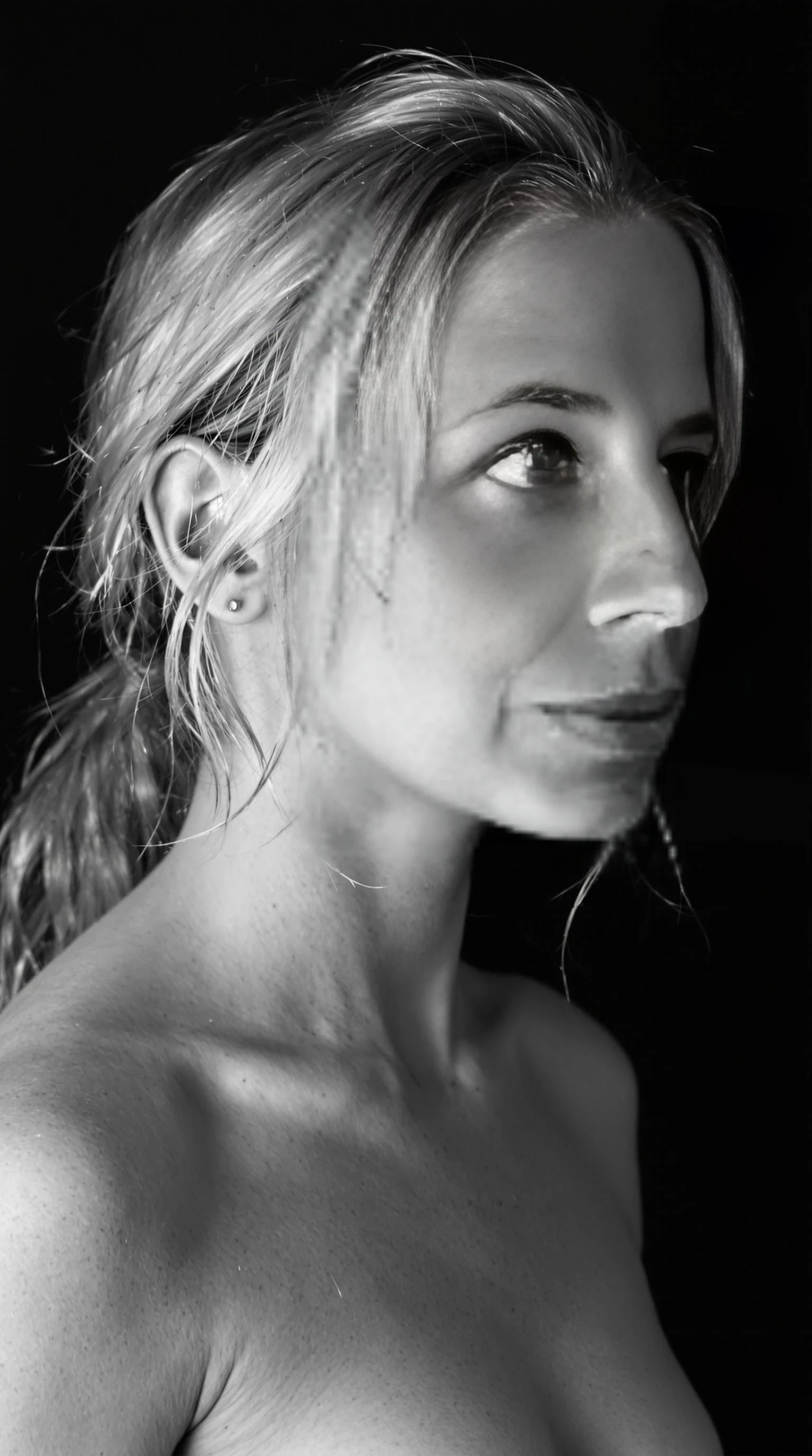 This is a black and white photograph of a young CSTMDL with fair skin and long, wavy blonde hair. She has a delicate, symmetrical face with high cheekbones and a slender neck. Her hair is pulled back, with some strands framing her face, creating a soft, ethereal look. She is wearing nothing, whole body exposed, she show her small imperfect breasts. The background is a solid, dark black, making her features and the details of her outfit stand out starkly. The lighting is soft and diffused, casting subtle shadows that highlight the contours of her face and the texture of her hair. The overall mood of the image is introspective and serene, emphasizing the subject's calm and thoughtful expression. The photograph captures a moment of quiet contemplation, with the subject gazing slightly to the right. The monochromatic color scheme adds a timeless and classic quality to the image.