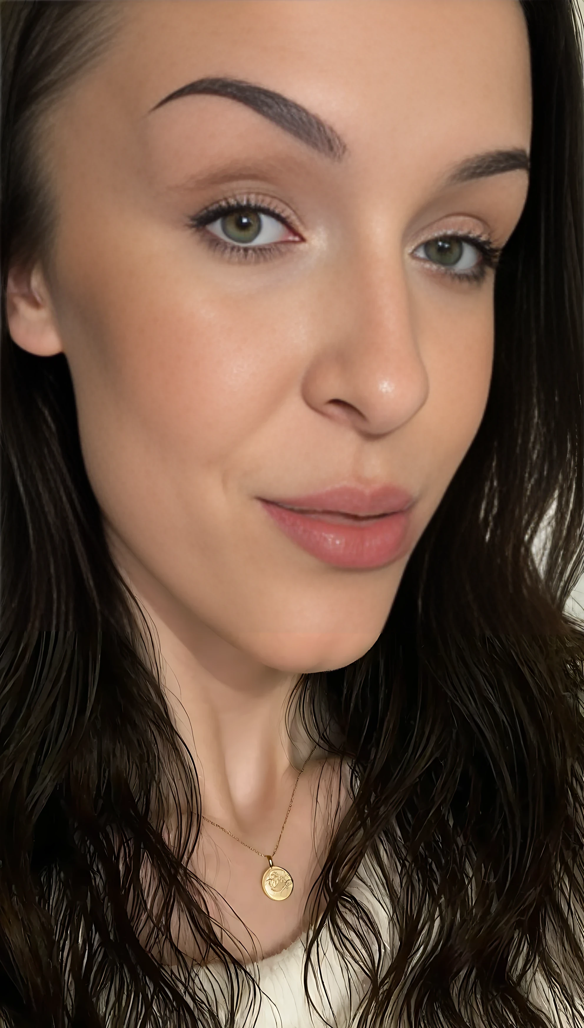 photo of Lena medium shot, selfie of a beautiful young woman with fair skin and smooth complexion, soft natural lighting. She has long, dark brown wavy hair, well-groomed thick eyebrows, almond-shaped hazel-green eyes with defined eyelashes, and subtle winged eyeliner. Her lips are full with a natural rosy tone, and she wears a soft matte nude lipstick. Minimal makeup look with a focus on natural beauty. She has a symmetrical face, slightly angled jawline, and a calm, confident expression. She is wearing a white fluffy sweater and a small golden pendant necklace. Neutral background, studio-quality photograph, photorealistic detail, 85mm lens, shallow depth of field.
