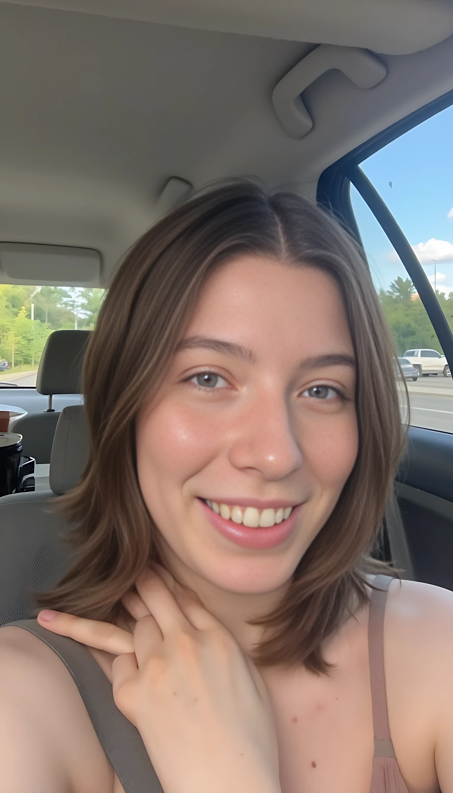 boring low quality snapchat photo circa 2015, A young woman with a warm smile, sitting topless in the driver's seat of a cozy car. She has soft, light brown hair cascading past her shoulders. The sunlight streams through the window, casting a gentle glow on her face. The interior of the car is neat and comfortable, with a coffee cup in the cup holder and a small plant on the dashboard. The background outside the window shows a scenic road lined with trees and blue skies