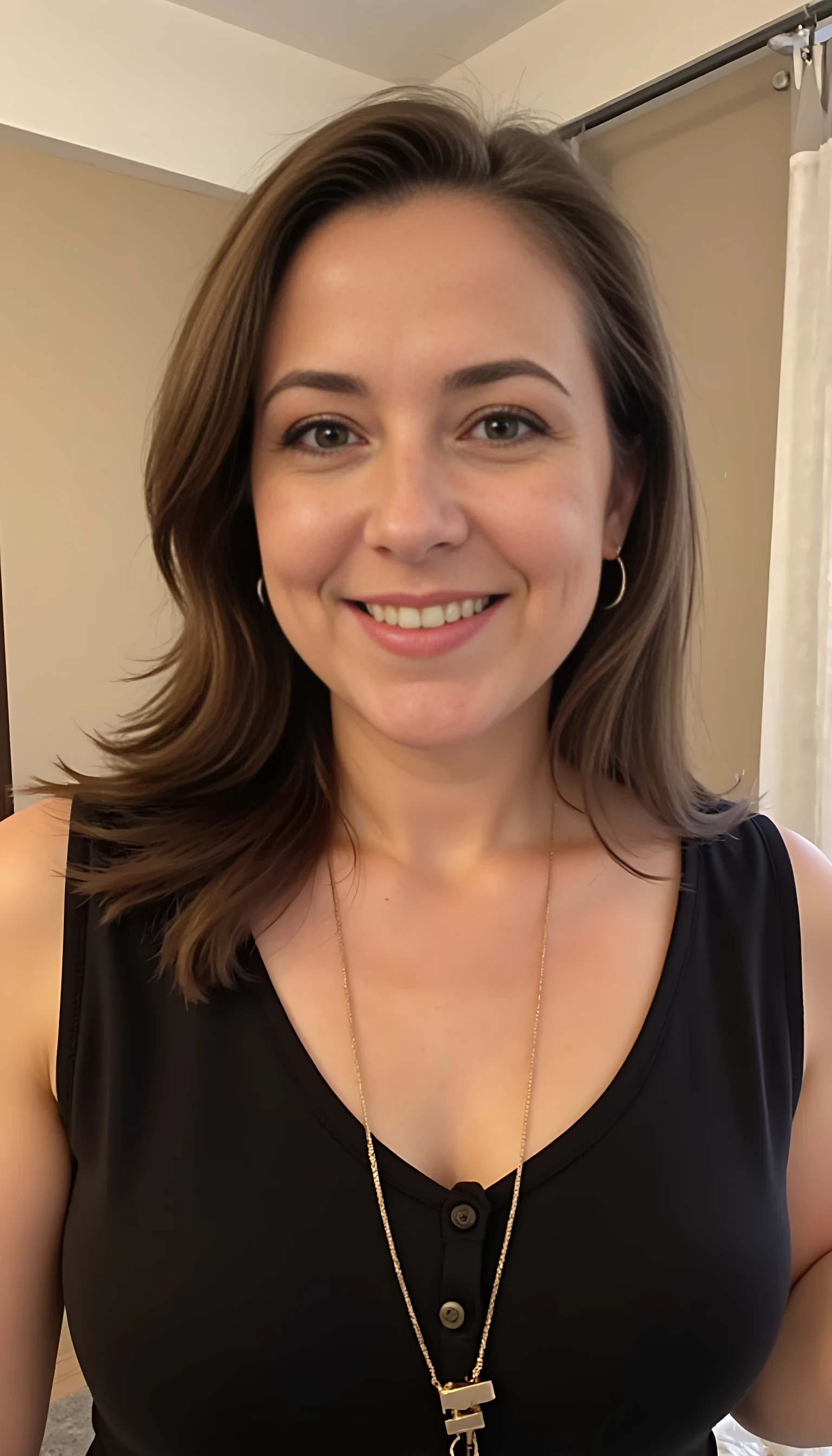 Photograph of a smiling CSTMDL wearing a black dress. She has small earrings and a gold necklace. Background is a beige wall with a silver curtain rod.