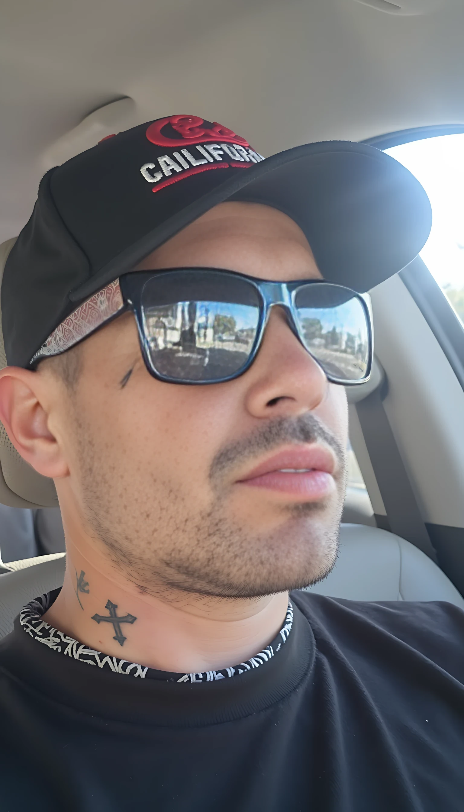 This is a photograph of a man sitting in a car, viewed from the side. He has a medium build and light skin tone, with a closely shaved head and facial hair. He is wearing a black baseball cap with a red logo and the text "CALIFORNIA" in white letters. The cap is tilted to the right, partially obscuring his face. His eyes are covered by large, reflective sunglasses with a black frame and a red and white paisley pattern on the lenses. The man's left ear is adorned with a small tattoo of a cross. He is wearing a black t-shirt with a white pattern on the collar.

In the background, the interior of the car can be seen, with a light-colored seat and the steering wheel visible. The windows show a blurred view of a bright, sunny day with trees and a building in the distance. The overall lighting in the photograph is bright and natural, indicating it was taken during the day. The man's expression is neutral, and his posture is relaxed. The image captures a casual, everyday moment.