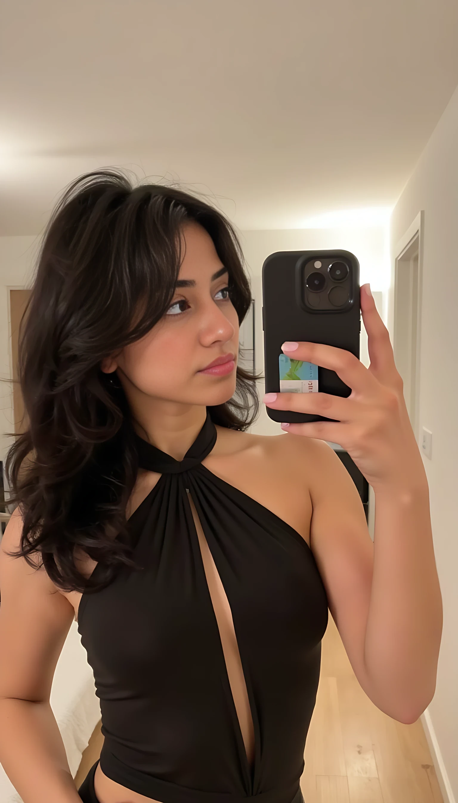 Portrait of a person taking a mirror selfie indoors, wearing a dark sleeveless halter top. The individual has long, wavy dark hair and is holding a phone with a card in the case, showing pastel-colored nails. The room has a modern aesthetic with light-colored wooden floors, a visible bed in the background, and soft lighting that gives a warm atmosphere, nude.