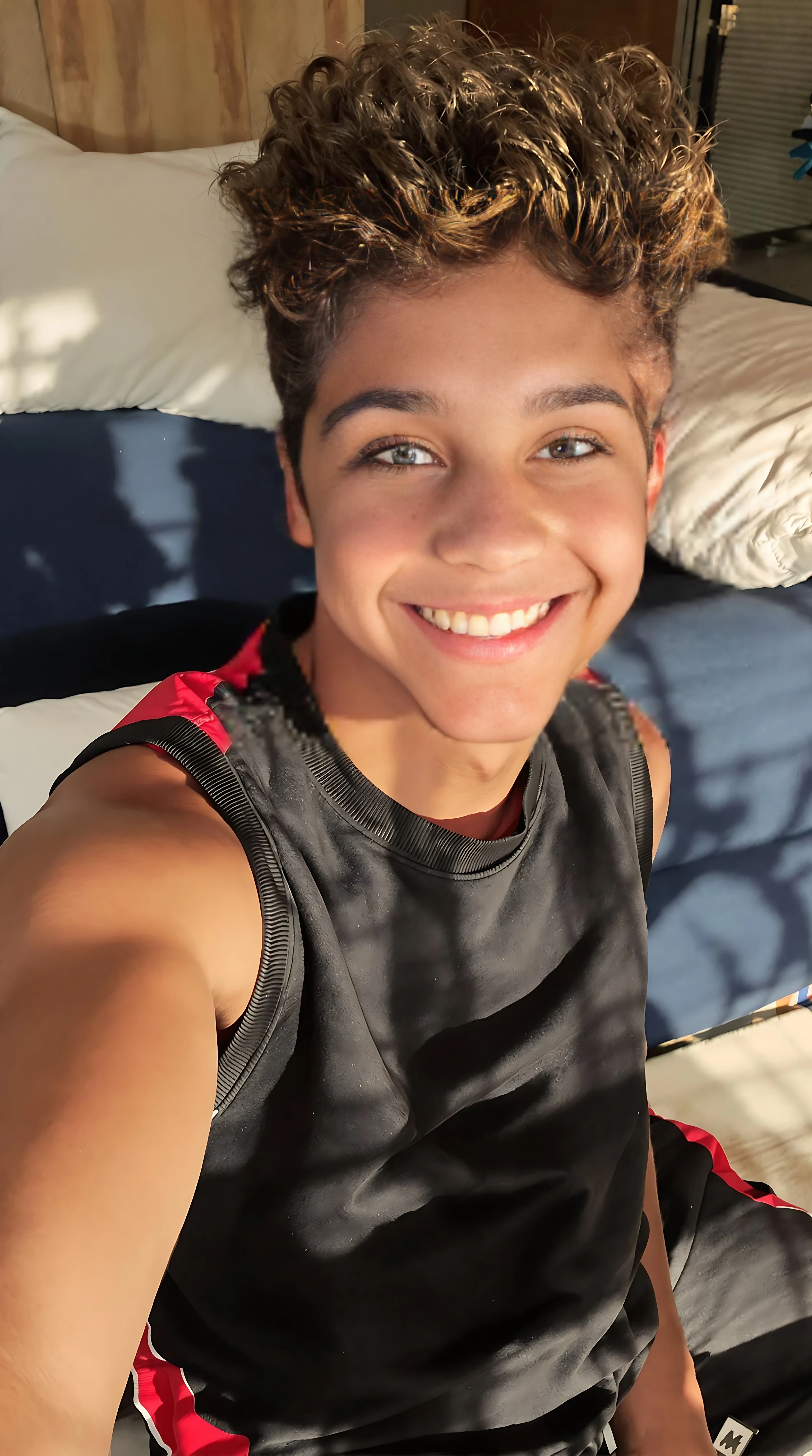 boring low quality snapchat photo circa 2015. A close up selfie style head shot photo of a 16-year-old boy, sitting on the couch, warmly lit in the late afternoon. The image is rich in detail, with warm tones and clear textures throughout. The boy has tanned skin, a medium, athletic build, brown curly hair and brown almond-shaped eyes. He is smiling subtly and warmly at the camera and wears a black and red sports jumper. The background features his lounge room and couch that is navy blue with white pillows. The overall composition is balanced, with the young boy looking comfortable. The details are vivid, with textures of clothing, skin, and background elements clearly visible, while the warm lighting creates a relaxed and joyful mood