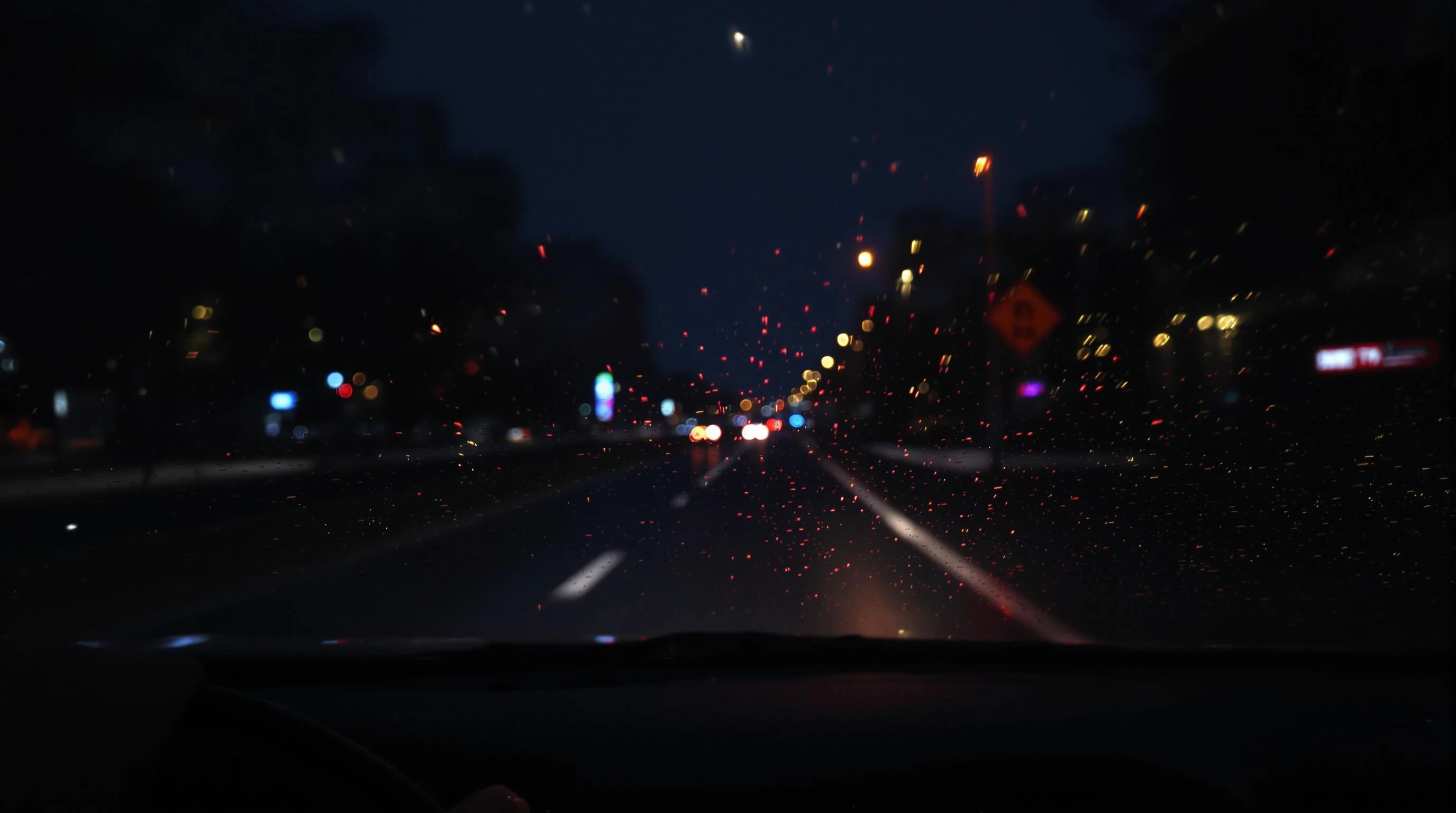 "A realistic nighttime scene viewed from inside a moving car, showing blurred city lights through a rain-covered windshield. The road is illuminated by soft neon reflections and distant headlights. The dashboard is visible in the foreground, with a pair of hands on the steering wheel, suggesting calm and focus. The mood is cinematic, introspective, and peaceful – ideal for a night drive. Ultra-realistic, moody lighting, 4K photo quality, shallow depth of field."