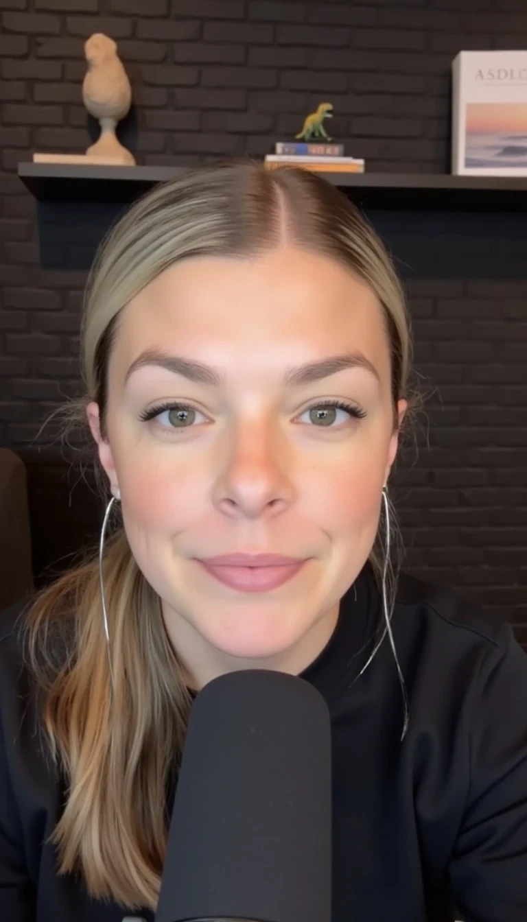 A woman with blonde hair pulled back, wearing a black shirt, is speaking into a microphone. The background features a dark brick wall and a shelf with books and decorative items, including a small dinosaur figure and a picture of a serene landscape on a book cover. The setting appears to be a cozy, modern podcast studio with soft lighting.