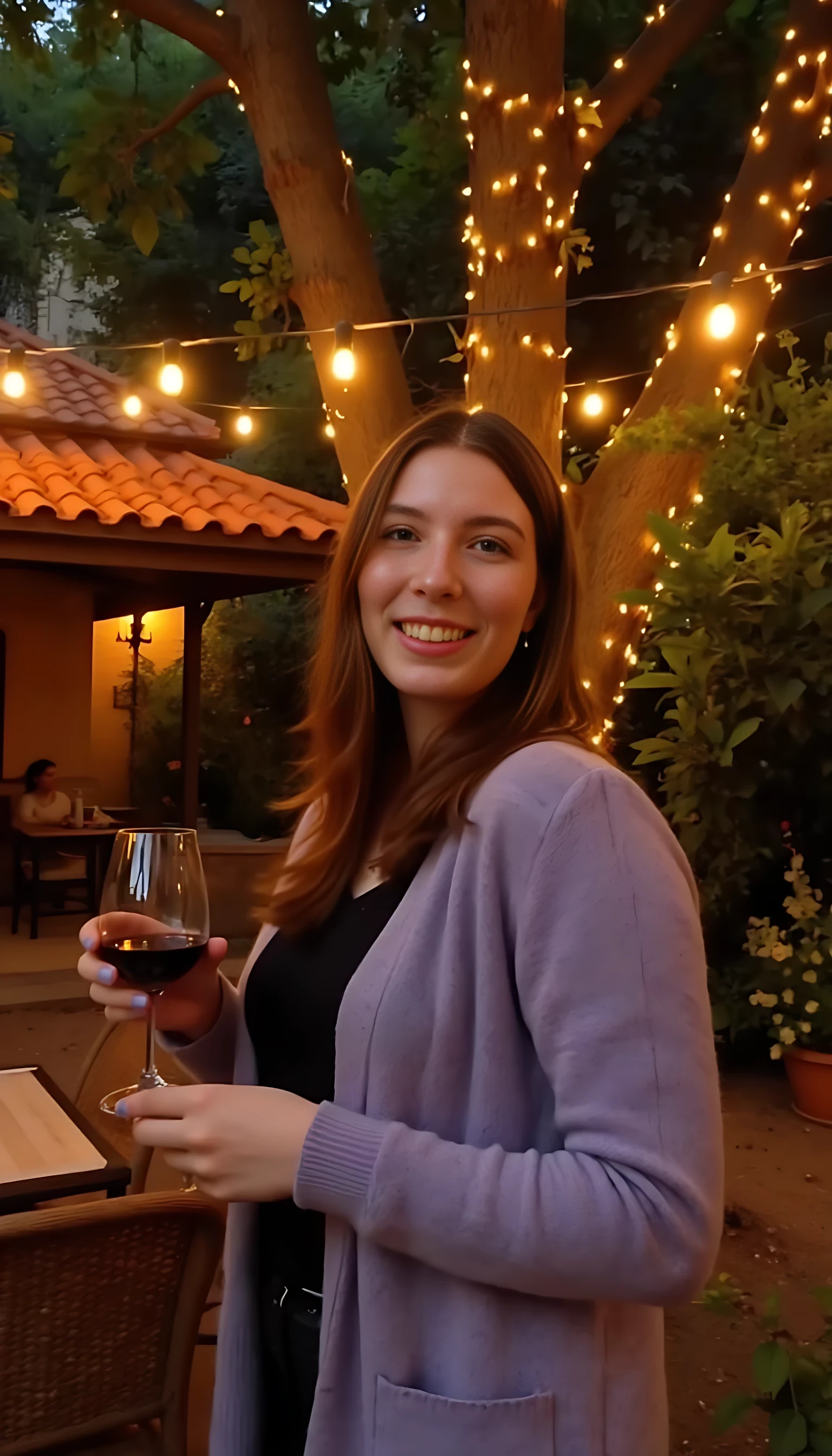 A full body shot photo of a woman standing outdoors at a cozy, warmly lit patio in the evening, with soft, glowing lights wrapped around a tree behind them creating a charming atmosphere. The image is rich in detail, with warm tones and clear textures throughout. The woman has fair skin, a slim build, and long reddish-blonde hair, also worn loose. She is smiling warmly at the camera, holding a glass of red wine in her right hand, and wears a soft-textured lavender cardigan over a black top. The background features a large tree with sparkling string lights wrapped around its trunk and branches, adding a magical touch to the scene. The setting suggests a private outdoor seating area, with elements like terracotta-tiled roofing, greenery, and rattan chairs contributing to a serene, inviting vibe. The overall composition is balanced, with the woman framed against the softly glowing tree, and the lighting enhances the warm, intimate feel of a quiet evening shared among friends. The details are vivid, with textures of clothing, skin, and background elements clearly visible, while the warm lighting creates a relaxed and joyful mood