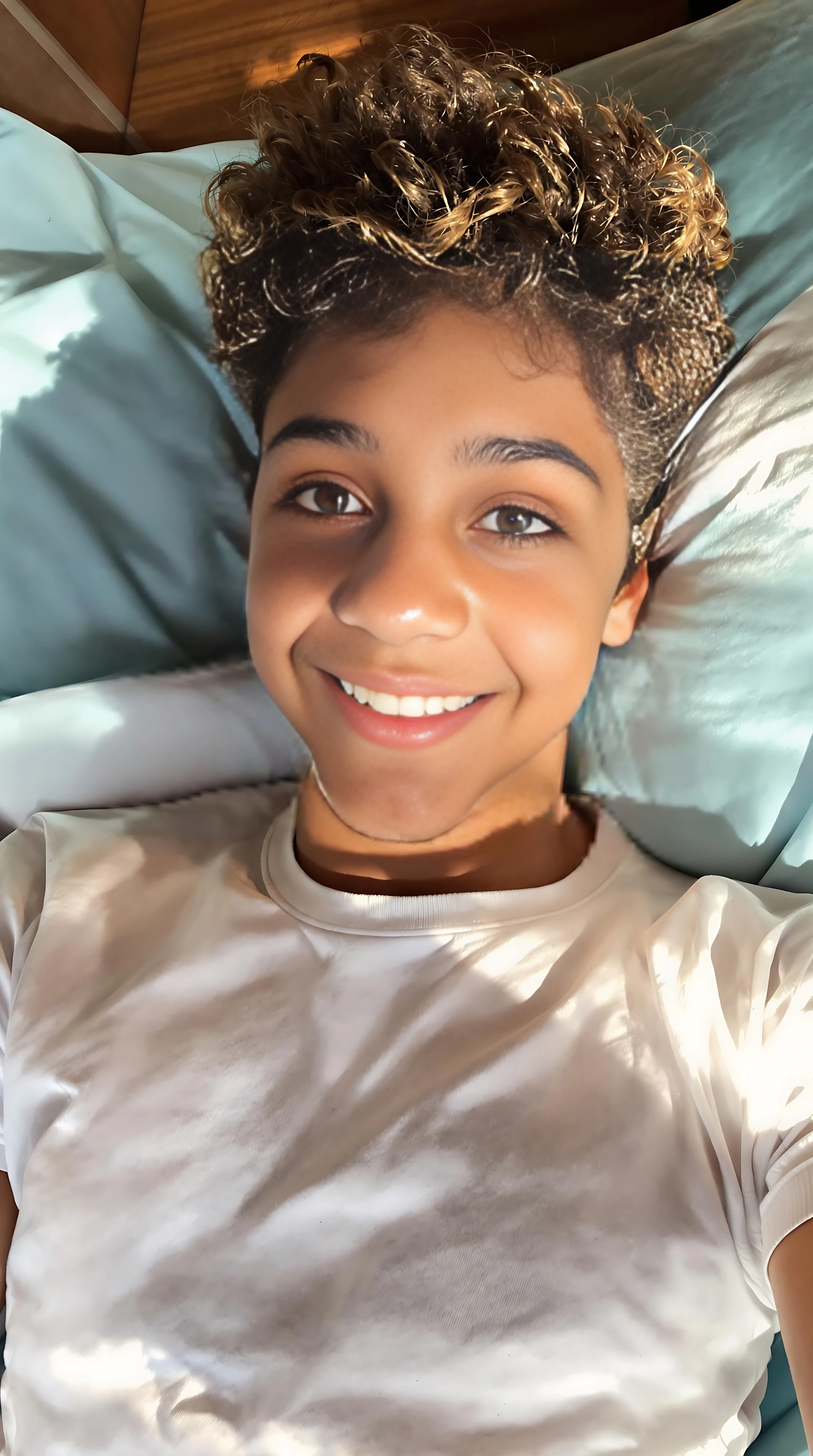 boring low quality snapchat photo circa 2015. A close up selfie style head shot photo of a 16-year-old boy, laying on his bed, warmly lit in the late afternoon. The image is rich in detail, with warm tones and clear textures throughout. The boy has tanned skin, a medium, athletic build, brown curly hair and brown almond-shaped eyes. He is smiling subtly and warmly at the camera and wears a white t-shirt. The background features his bedding, light blue sheets and pillows. The overall composition is balanced, with the young boy looking comfortable and his face is lit up from the window, and the afternoon lighting evoking a summery feel. The details are vivid, with textures of clothing, skin, and background elements clearly visible, while the warm lighting creates a relaxed and joyful mood