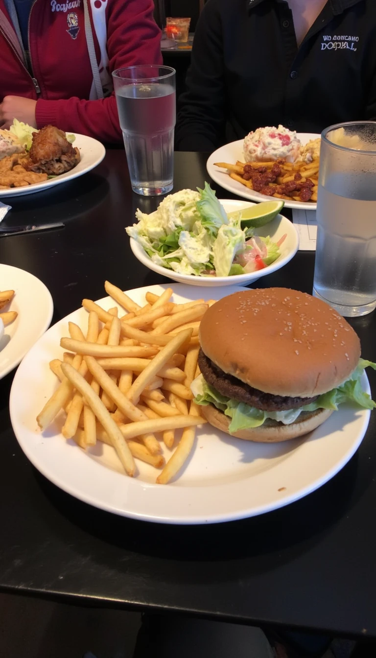 boring low quality snapchat photo circa 2015, this picture is being taken in a restaurant. The picture is of a Burger and fries in on one plate and a bowl of ceaser salad next to it. There are other food dishes and drink glasses on the table as well. The food is in white dishes on a black table. 