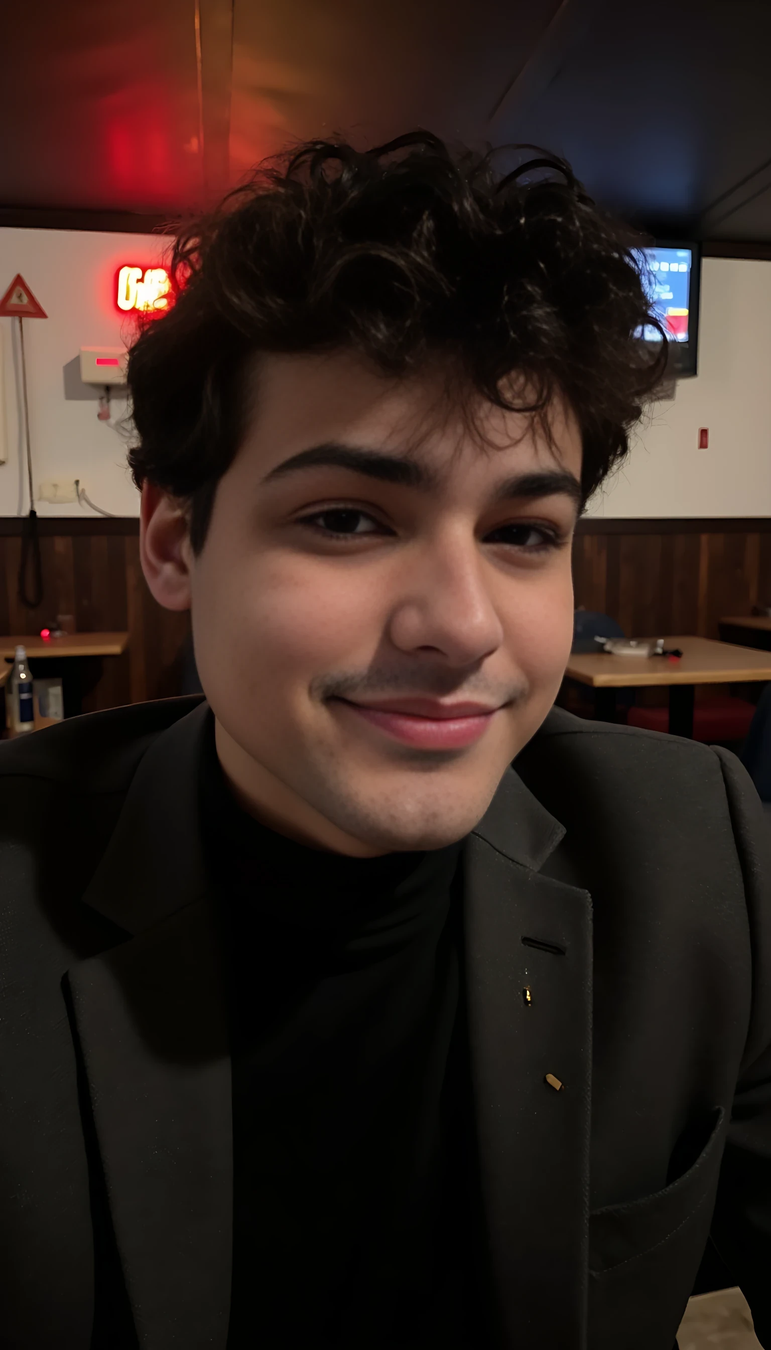 Boring low-quality Snapchat photo of a handsome 20-year-old man smiling at night, CSTMDL style. He's in a retro-chic bar with dim lighting and dark wood interior. Wearing a black turtleneck and gray wool blazer, subtle gold ring, and tousled hair. Cinematic mood, relaxed confident pose, looks like a spontaneous low-res phone picture with light grain and soft lens blur.