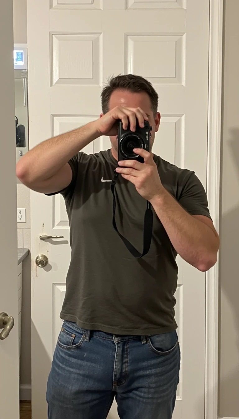 Picture of a muscular man in late 40s CSTMDL with brown hair. he is wearing a tshirt and jeans and is facing the picture while holding a camera up as if he is taking a picture. The camera is covering his face