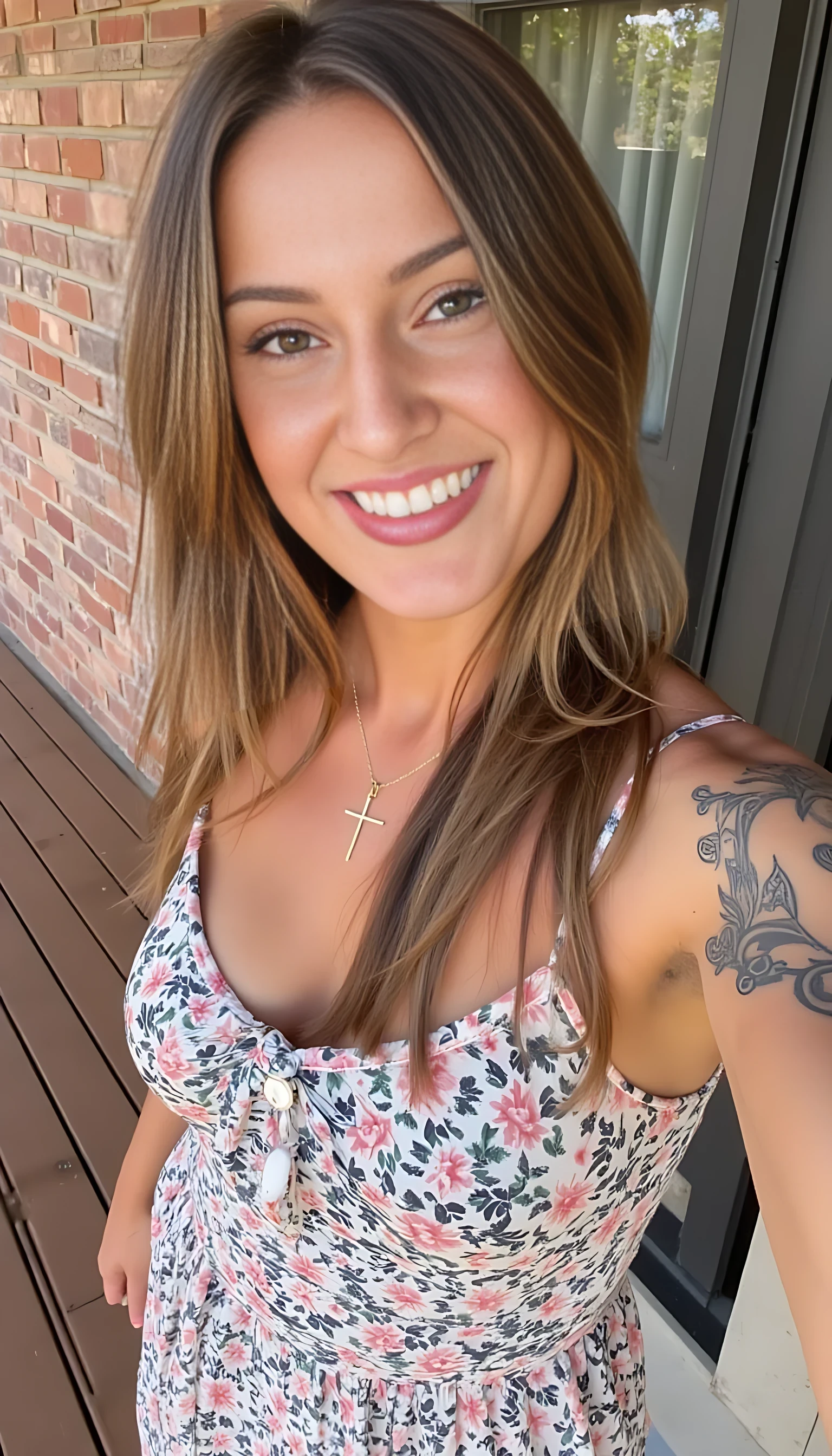 A confident woman taking a selfie on a wooden deck, smiling warmly. She has long, light brown hair and wears subtle makeup. Her floral-patterned dress is sleeveless, displaying intricate tattoos on her arms. A gold necklace with a cross pendant adorns her neckline. The background features a brick wall and a partially visible window with a curtain.
