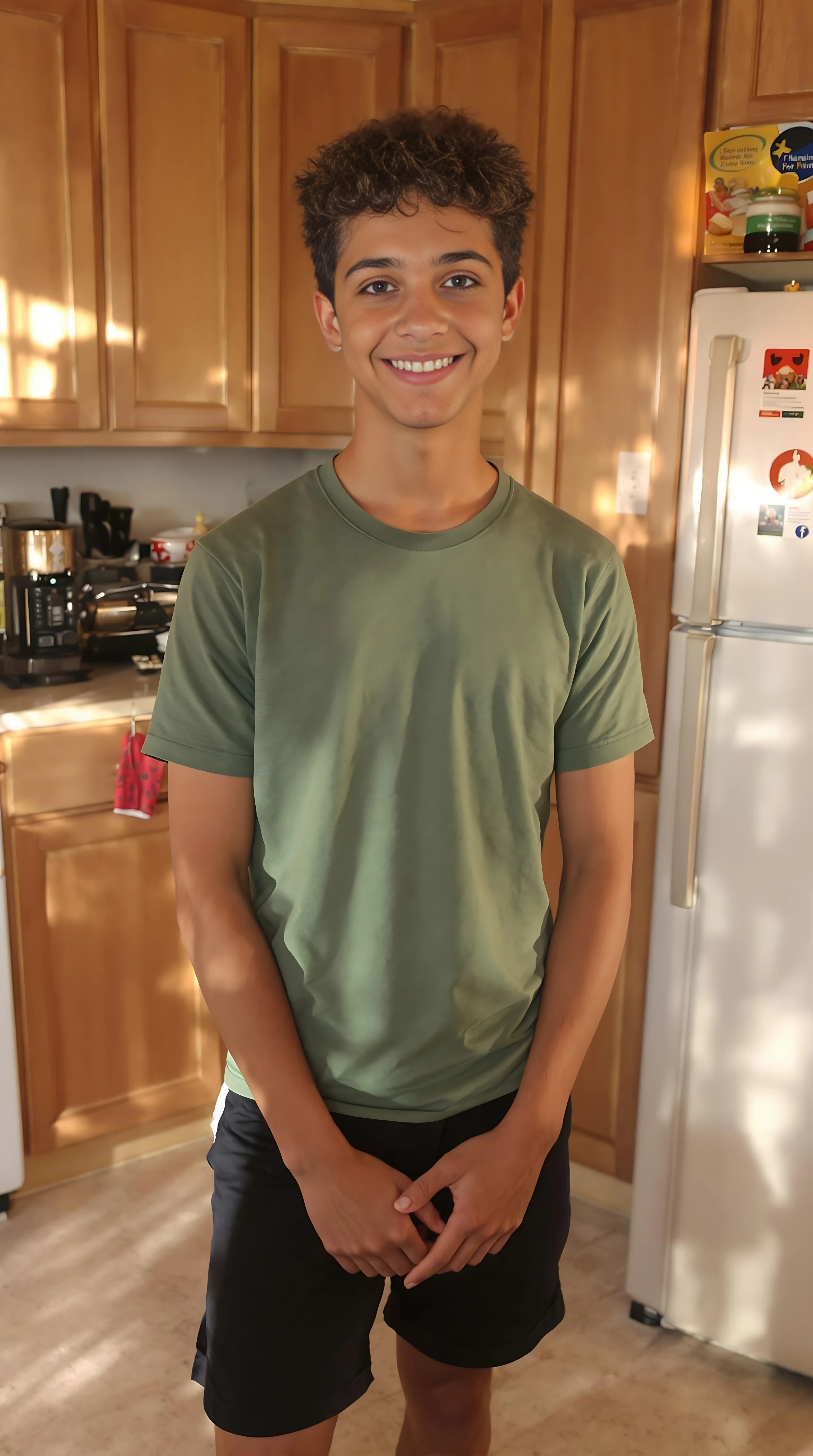 boring low quality snapchat photo circa 2015. A selfie photo of a 16-year-old boy, standing in his family home kitchen, warmly lit in the late afternoon. The image is rich in detail, with warm tones and clear textures throughout. The boy has tanned skin, a medium, athletic build, brown curly hair and brown almond-shaped eyes. He is smiling subtly and warmly at the camera, he's wearing a green t-shirt and black shorts.