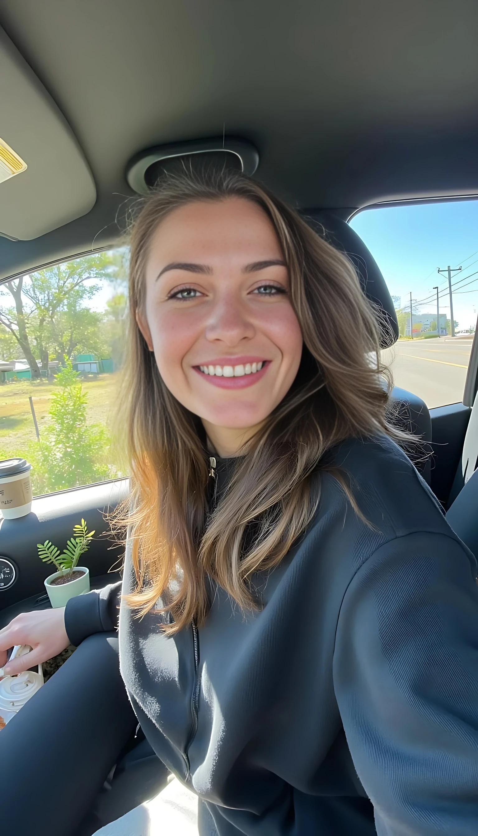 boring low quality snapchat photo circa 2015, A young woman with a warm smile, sitting in the driver's seat of a cozy car. She has soft, wavy brown hair cascading past her shoulders. The sunlight streams through the window, casting a gentle glow on her face. The interior of the car is neat and comfortable, with a coffee cup in the cup holder and a small plant on the dashboard. The background outside the window shows a scenic road lined with trees and blue skies