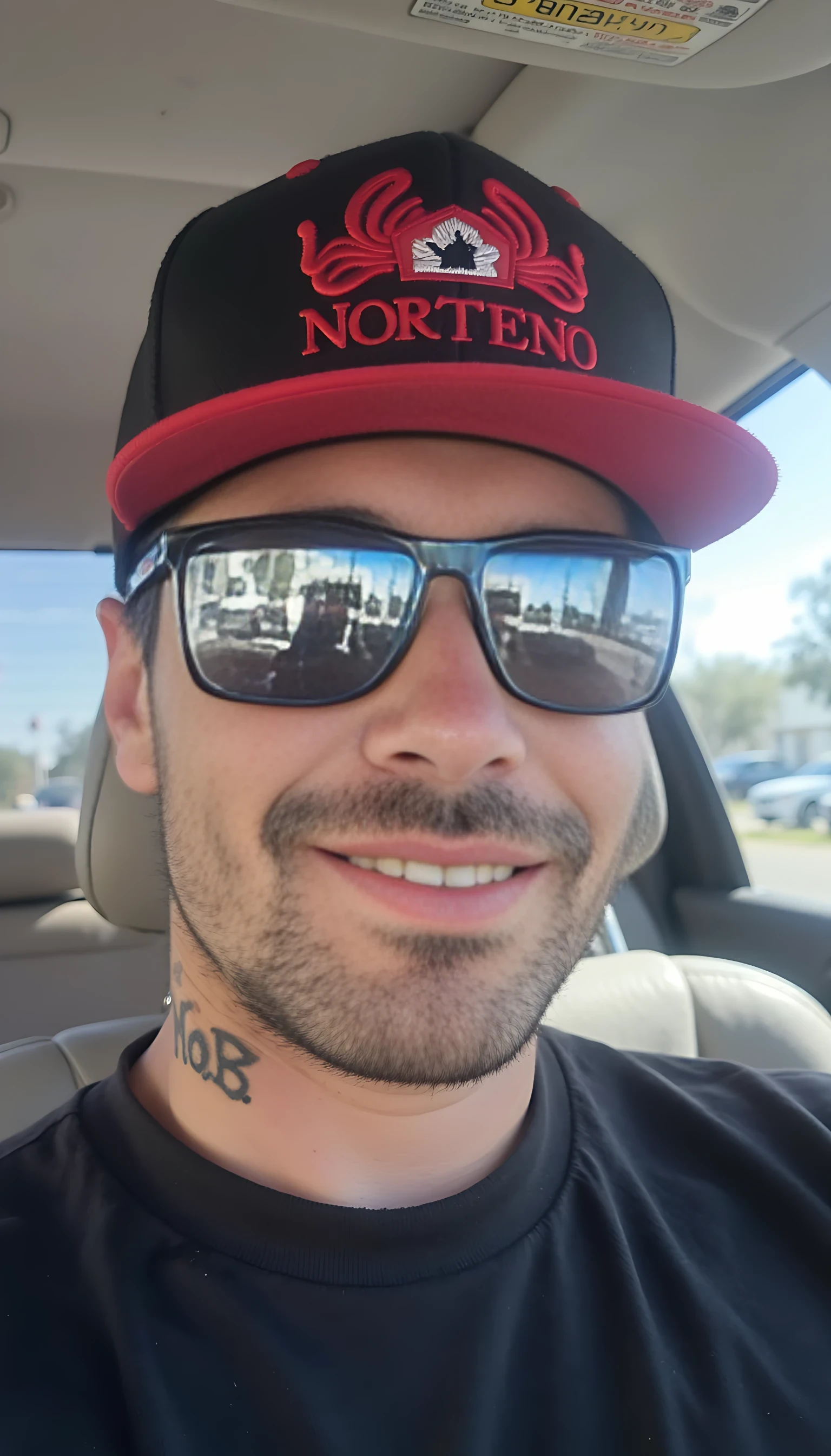 This is a photograph of a man sitting in a car, viewed from the side. He has a medium build and light skin tone, with a closely shaved head and facial hair. He is wearing a black snapback cap with a red bill and logo and the text "NORTENO" in red letters. The cap is tilted to the right, with a straight bill partially obscuring his face. His eyes are covered by large, reflective sunglasses with a black frame and a red and white paisley pattern on the lenses. The man's left ear is adorned with a small tattoo that reads "M.O.B. in neatly written old english lettering. He is wearing a black t-shirt with a large real diamond chain on, and a gold grill in his mouth. Smiling for the camera

In the background, the interior of the car can be seen, with a light-colored seat and the steering wheel visible. The windows show a blurred view of a bright, sunny day with trees and a building in the distance. The overall lighting in the photograph is bright and natural, indicating it was taken during the day. The man's expression is neutral, and his posture is relaxed. The image captures a casual, everyday moment.