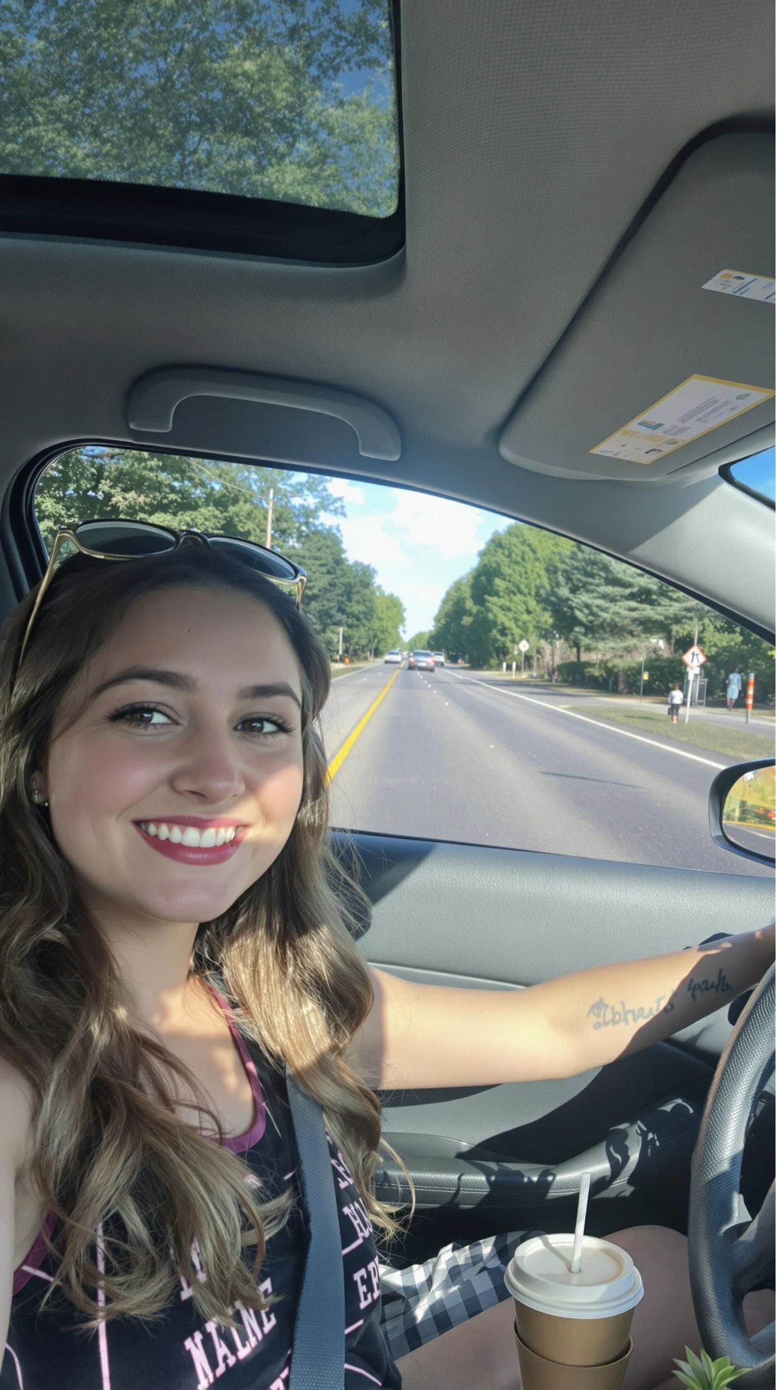 boring low quality snapchat photo circa 2015, A young woman with a warm smile, sitting in the driver's seat of a cozy car. She has soft, wavy brown hair cascading past her shoulders. The sunlight streams through the window, casting a gentle glow on her face. The interior of the car is neat and comfortable, with a coffee cup in the cup holder and a small plant on the dashboard. The background outside the window shows a scenic road lined with trees and blue skies