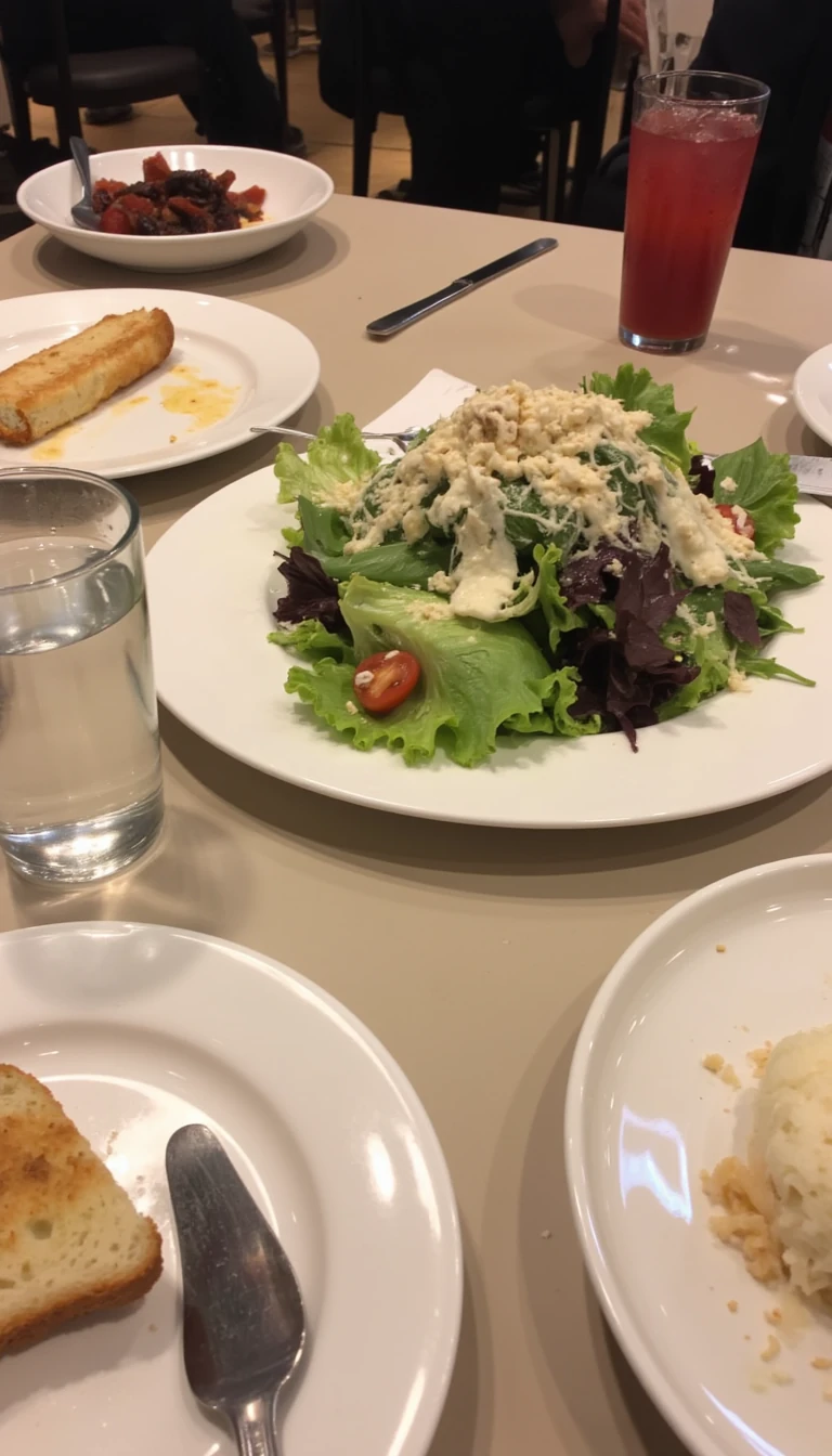 boring low quality snapchat photo circa 2015, this picture is being taken in a restaurant. The picture is of large salad with a glass of water next to it. There are other food dishes and drink glasses on the table as well.  All the food dishes are white.