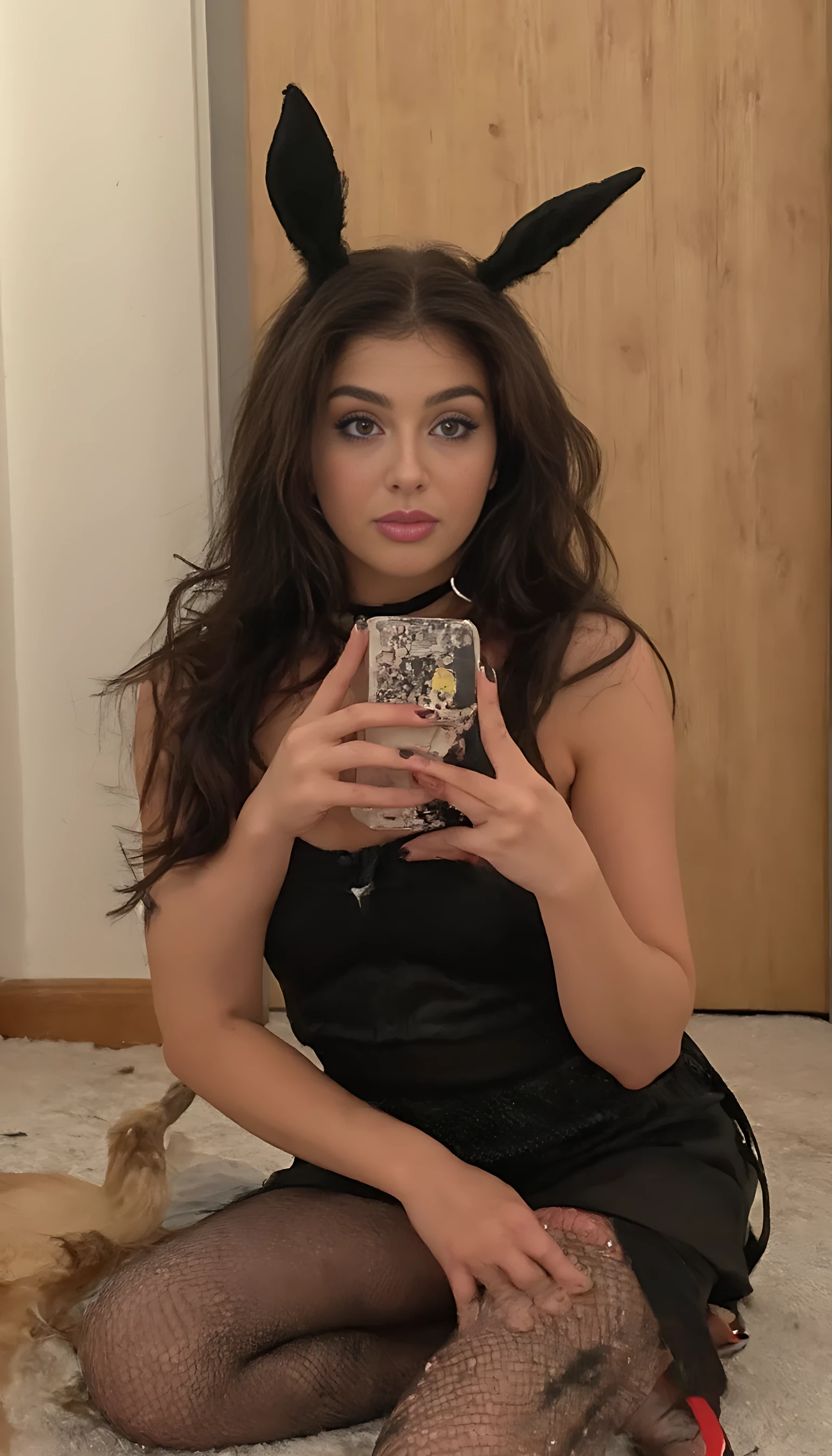 CSTMDL, 19 year old woman, 160cm tall, C cups, wavy dark brown hair framing face, softly arched full brows, hazel-brown eyes, long curled lashes, subtle bronze eyeshadow, gentle highlight on brow bone, straight slender nose with soft bridge, full heart-shaped lips with glossy rose tint, natural pink blush on apples of cheeks, faint freckles across nose, defined cheekbones with subtle contour, soft jawline tapering to delicate chin, slight chin dimple, natural skin texture with minimal pores, warm soft glow, high-resolution facial detail, The scene captures a confident female character posing playfully in a stylish costume reminiscent of a classic bunny outfit. She is seated on a soft, neutral-toned carpet, providing a cozy feel to the room, which features a softly lit backdrop emphasizing warmth., the character has long, flowing hair, partially draped over one shoulder, which adds to her alluring appearance. Her facial features are highlighted by her playful expression, showcasing a subtle smile and inviting gaze as she holds a smartphone for a selfie, enhancing the modern vibe of the image. She wears a black bodysuit that hugs her figure, accentuating her curves, complemented by sheer fishnet stockings that add an edgy element to her look. The outfit is topped with iconic bunny ears, contributing to a playful theme, and her footwear consists of striking red stilettos that add a pop of color and sophistication. The composition centers around her, drawing the viewer's attention to her posture, which is both relaxed and inviting. Soft, diffused lighting creates gentle shadows that accentuate her profile and the textures of her outfit, while the plain wall and door in the background keep the focus on her. 
