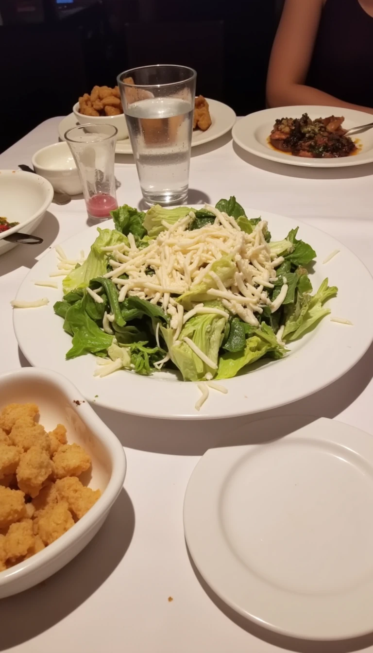 boring low quality snapchat photo circa 2015, this picture is being taken in a restaurant. The picture is of large caesar salad with a glass of water next to it. There are other food dishes and drink glasses on the table as well.  All the food dishes are white.