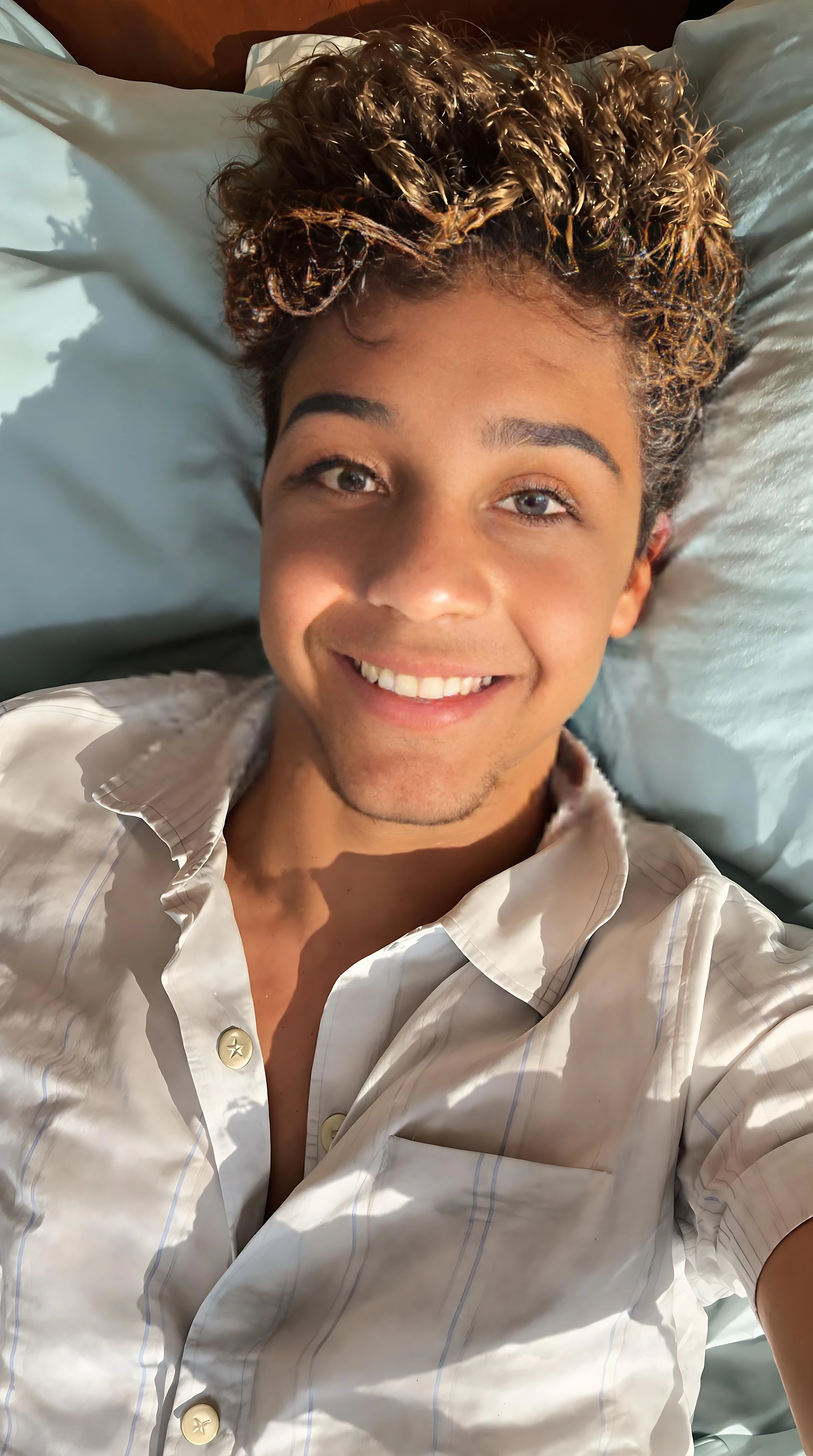boring low quality snapchat photo circa 2015. A close up selfie style head shot photo of a 16-year-old boy, laying on his bed, warmly lit in the late afternoon. The image is rich in detail, with warm tones and clear textures throughout. The boy has tanned skin, a medium, athletic build, brown curly hair and brown almond-shaped eyes. He is smiling subtly and warmly at the camera and wears a pj shirt. The background features his bedding, light blue sheets and pillows. The overall composition is balanced, with the young boy looking comfortable. The details are vivid, with textures of clothing, skin, and background elements clearly visible, while the warm lighting creates a relaxed and joyful mood