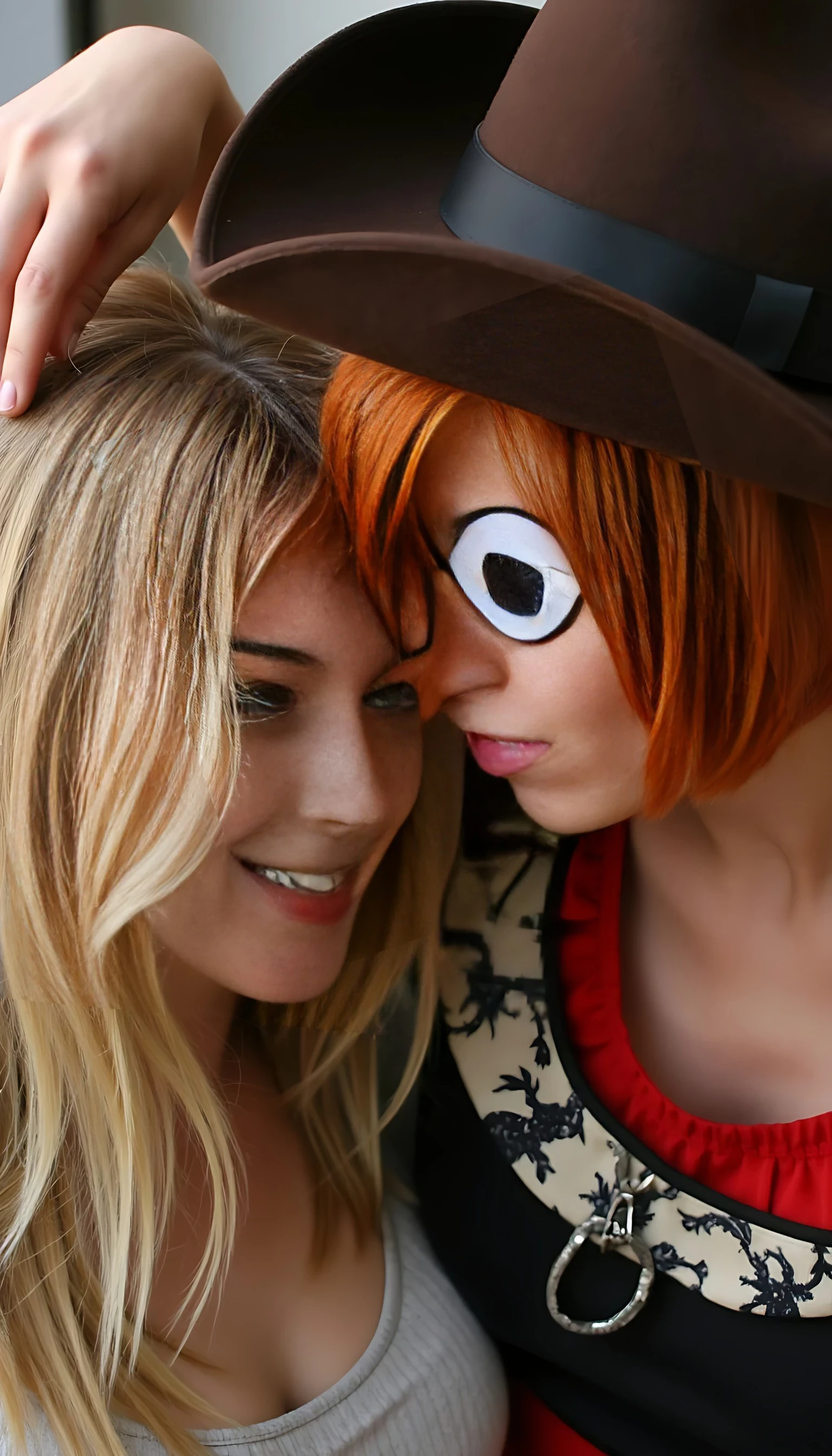 A short pixie style natural red head with an eye patch over her left eye wearing a brown cowboy hat is playing with a blonde woman’s long blonde hair. The blonde is enjoying it. Highly realistic photo. 