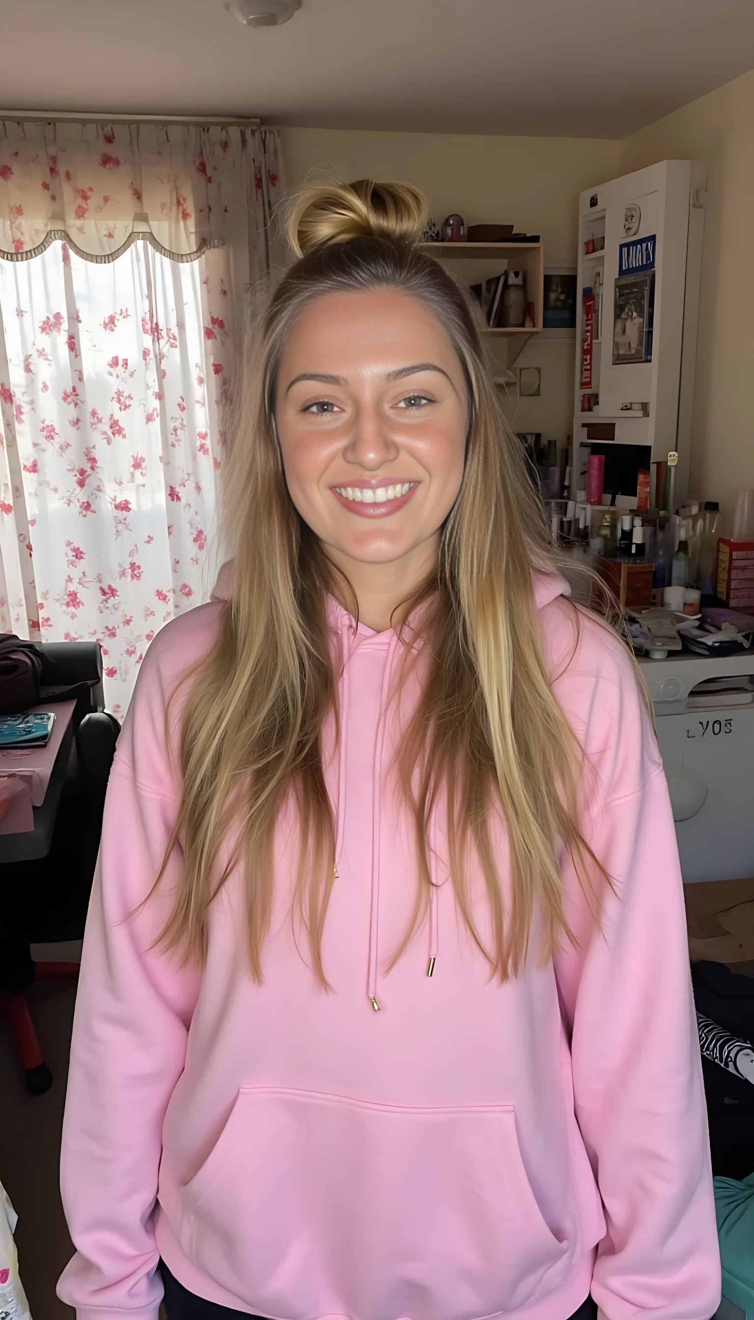 A young woman with long, highlighted hair smiles confidently in a cozy bedroom setting. Her hair is styled in a topknot with loose waves cascading down. She is wearing a pink hoodie draped over her shoulders and black pants. The room features floral patterned curtains and various personal items, creating a warm and lived-in atmosphere.