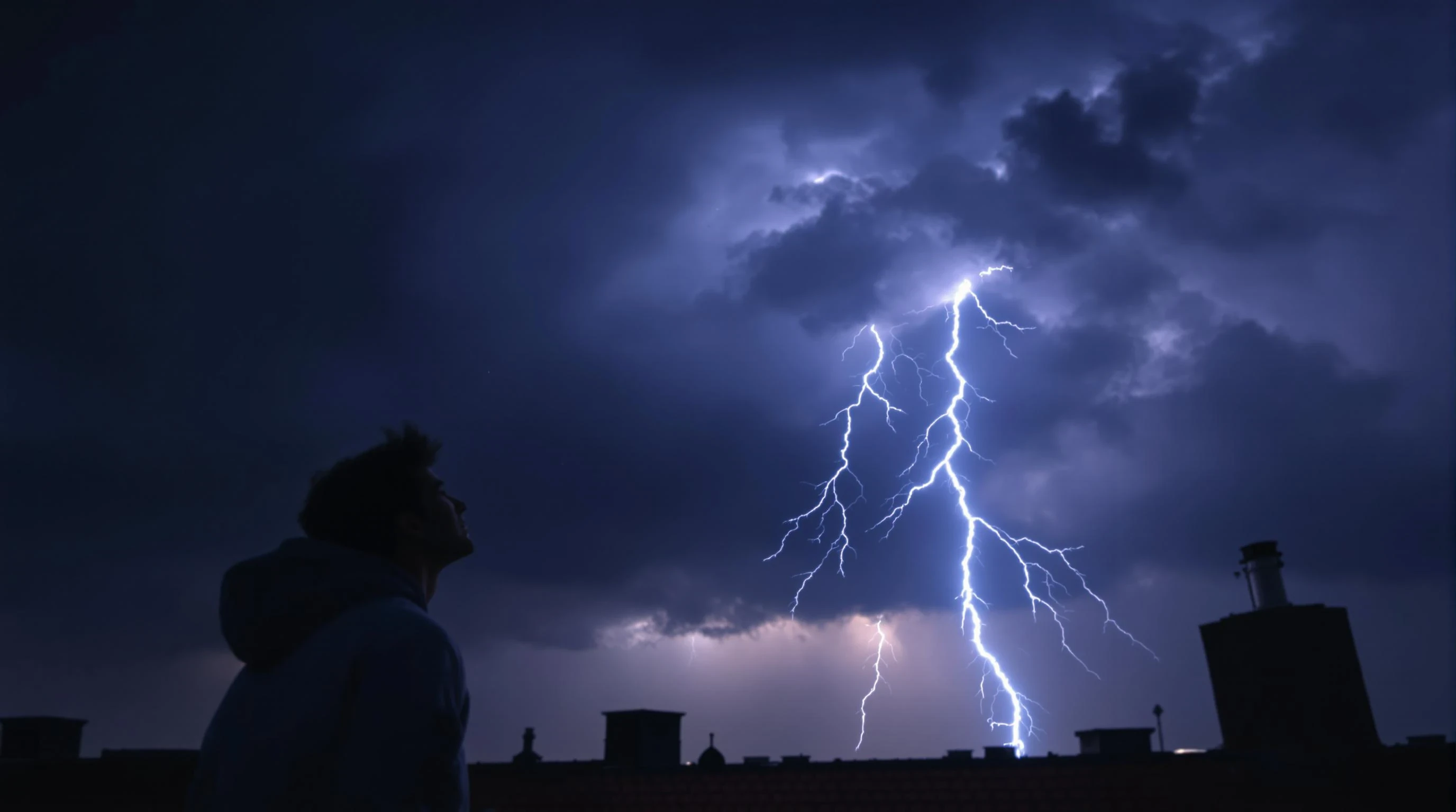 Ultra-realistic cinematic shot of a young man standing on a rooftop at night, looking up at a dramatic sky. In the clouds above, a massive lightning bolt strikes toward the ground, casting a blue-white glow over the scene. His face is lit by the lightning, wearing a casual hoodie, wind blowing slightly. Photorealistic, 8k, high detail, moody atmosphere, space in the center for a circular logo overlay.

