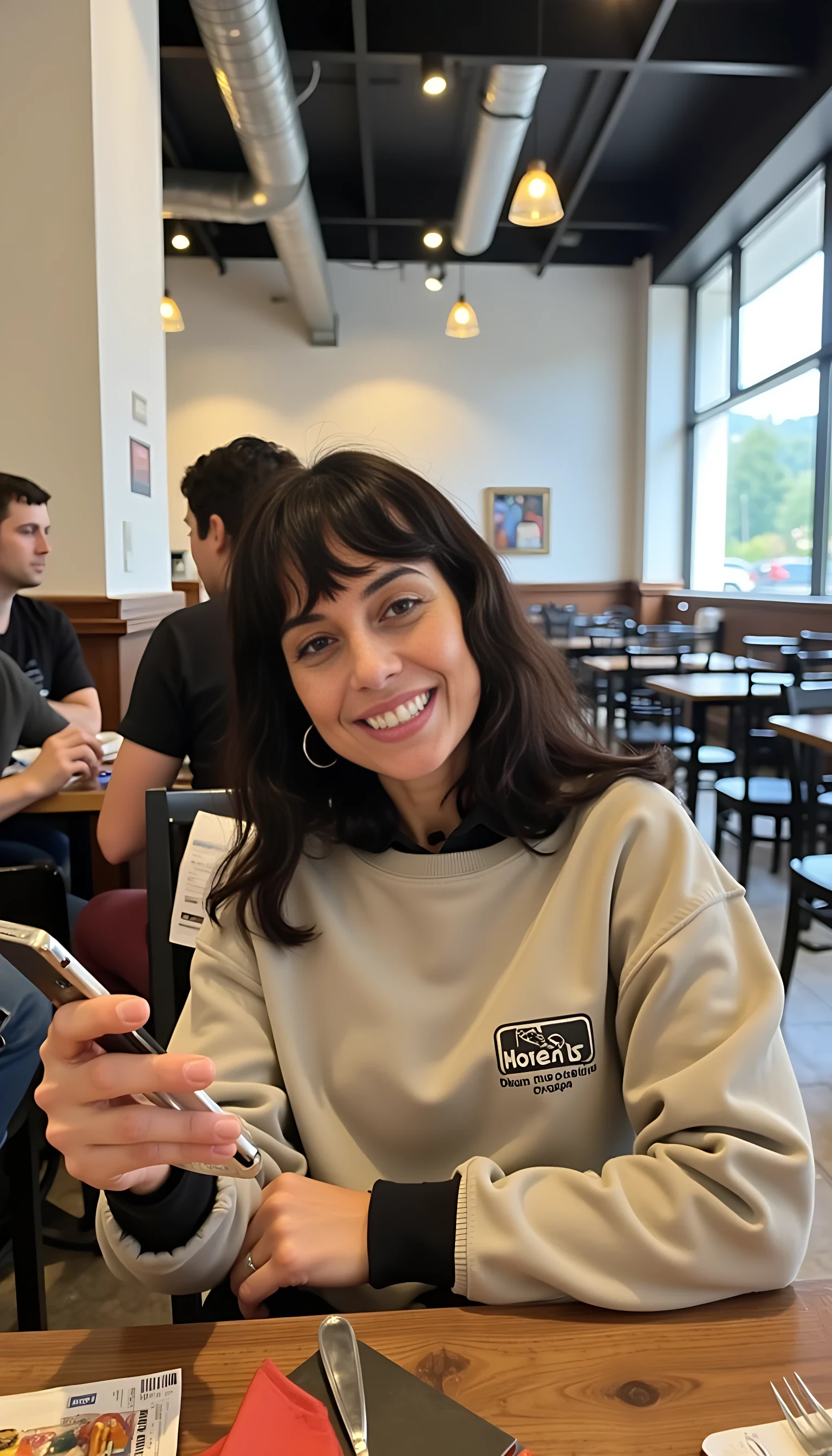iphone portrait of CSTMDL woman younger Bob length hair hair makeup bangs cute smile brown eyes and sitting in cafe holding phone in busy restaurant wearing hoodie says “wizard”  mountains background shopping bags 
