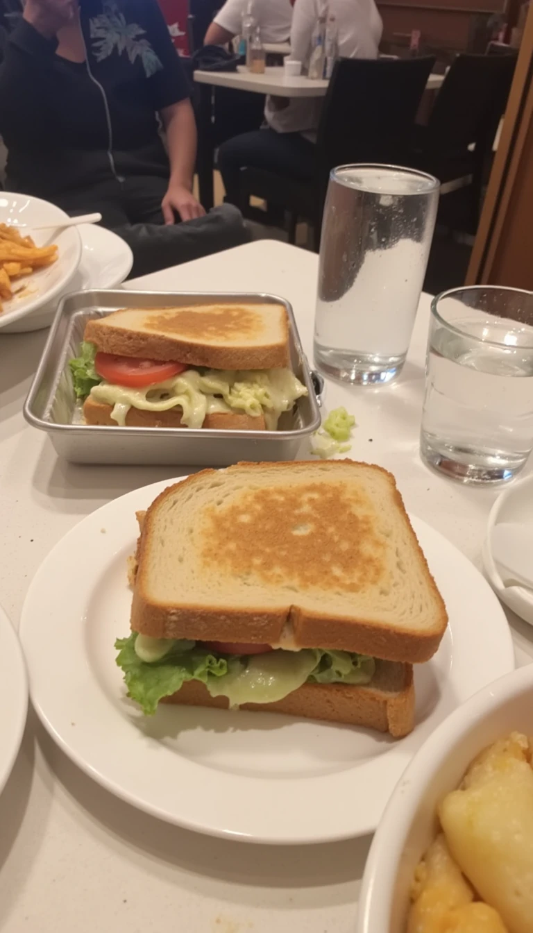 boring low quality snapchat photo circa 2015, this picture is being taken in a restaurant. The picture is of sandwich with a glass of water next to it. There are other food dishes and drink glasses on the table as well.  All the food dishes are white.