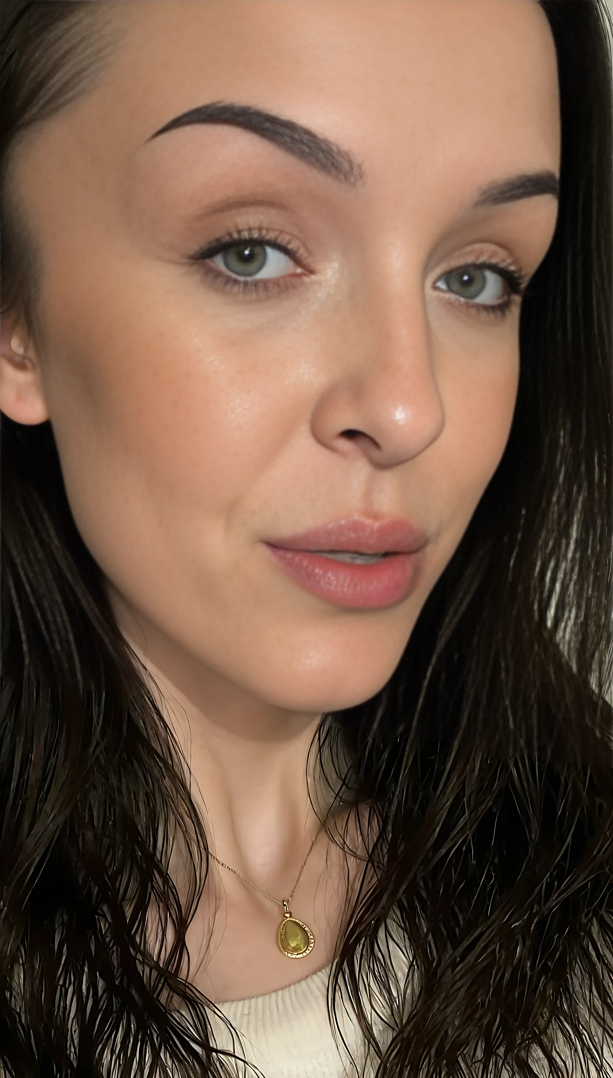 photo of Lena medium Close-up portrait of a beautiful young woman with fair skin and smooth complexion, soft natural lighting. She has long, dark brown wavy hair, well-groomed thick eyebrows, almond-shaped hazel-green eyes with defined eyelashes, and subtle winged eyeliner. Her lips are full with a natural rosy tone, and she wears a soft matte nude lipstick. Minimal makeup look with a focus on natural beauty. She has a symmetrical face, slightly angled jawline, and a calm, confident expression. She is wearing a white fluffy sweater and a small golden pendant necklace. Neutral background, studio-quality photograph, photorealistic detail, 85mm lens, shallow depth of field.
