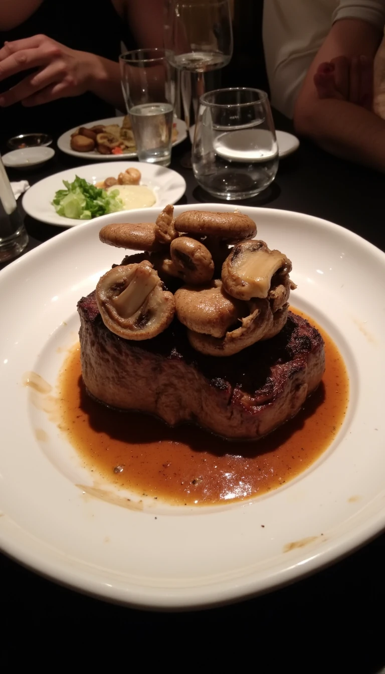 boring low quality snapchat photo circa 2015, this picture is being taken in a fancy restaurant. the picture is of a steak with mushroom's on top. The food is in a white dish on a black table. 
