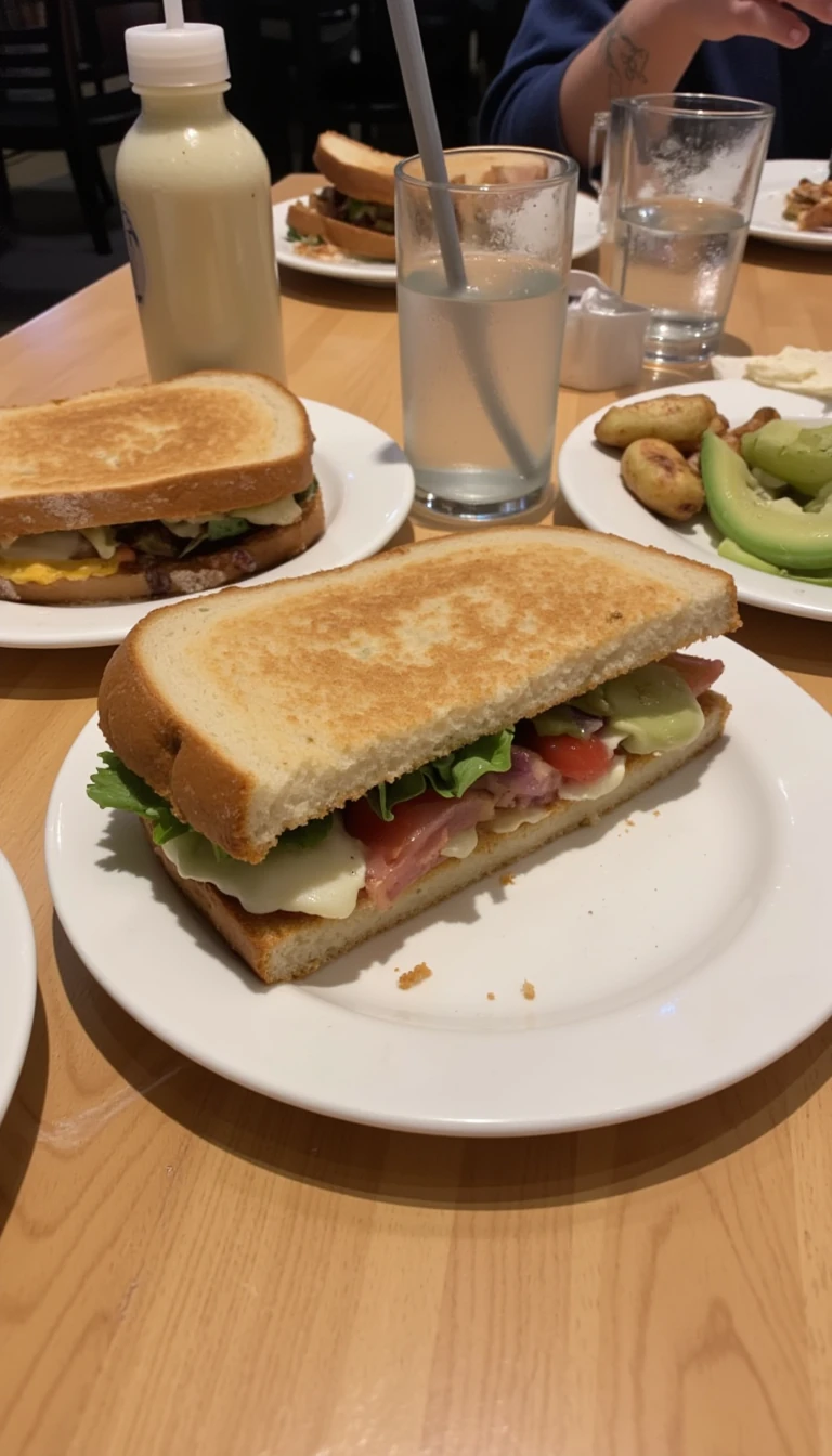 boring low quality snapchat photo circa 2015, this picture is being taken in a restaurant. The picture is of sandwich with a glass of water next to it. There are other food dishes and drink glasses on the table as well.  All the food dishes are white.