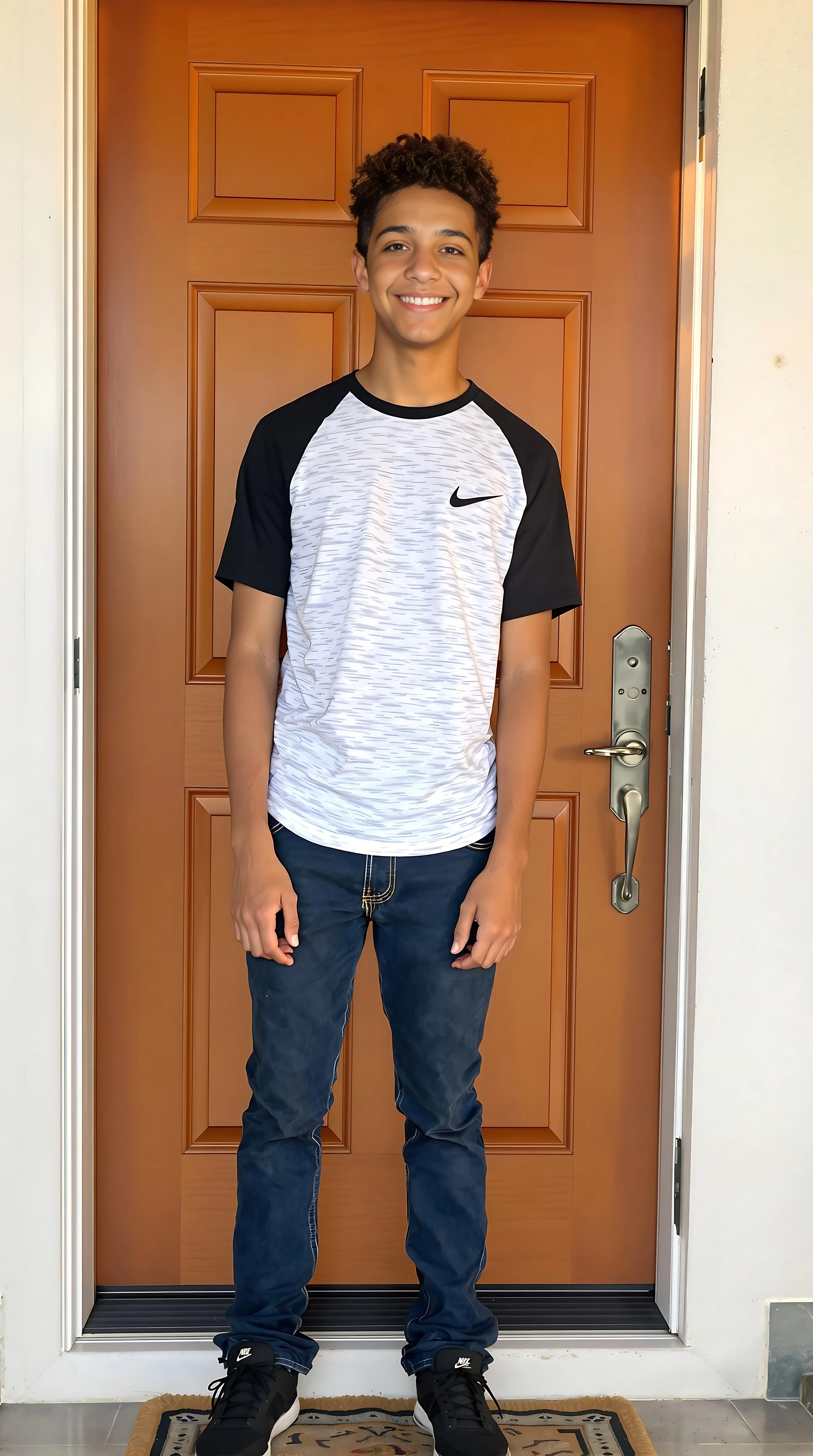 boring low quality snapchat photo circa 2015. A full body shot photo of a 16-year-old boy, standing by his front door in his home, warmly lit in the late afternoon. The image is rich in detail, with warm tones and clear textures throughout. The boy has tanned skin, a medium, athletic build, brown curly hair and brown almond-shaped eyes. He is smiling subtly and warmly away from the camera as if someone is standing in front of him out of frame. He wears a black and white t-shirt, blue jeans and black Nike runners. The background features his home front door as he stand in the doorway. The overall composition is balanced, with the young boy looking comfortable. The details are vivid, with textures of clothing, skin, and background elements clearly visible, while the warm lighting creates a relaxed and joyful mood